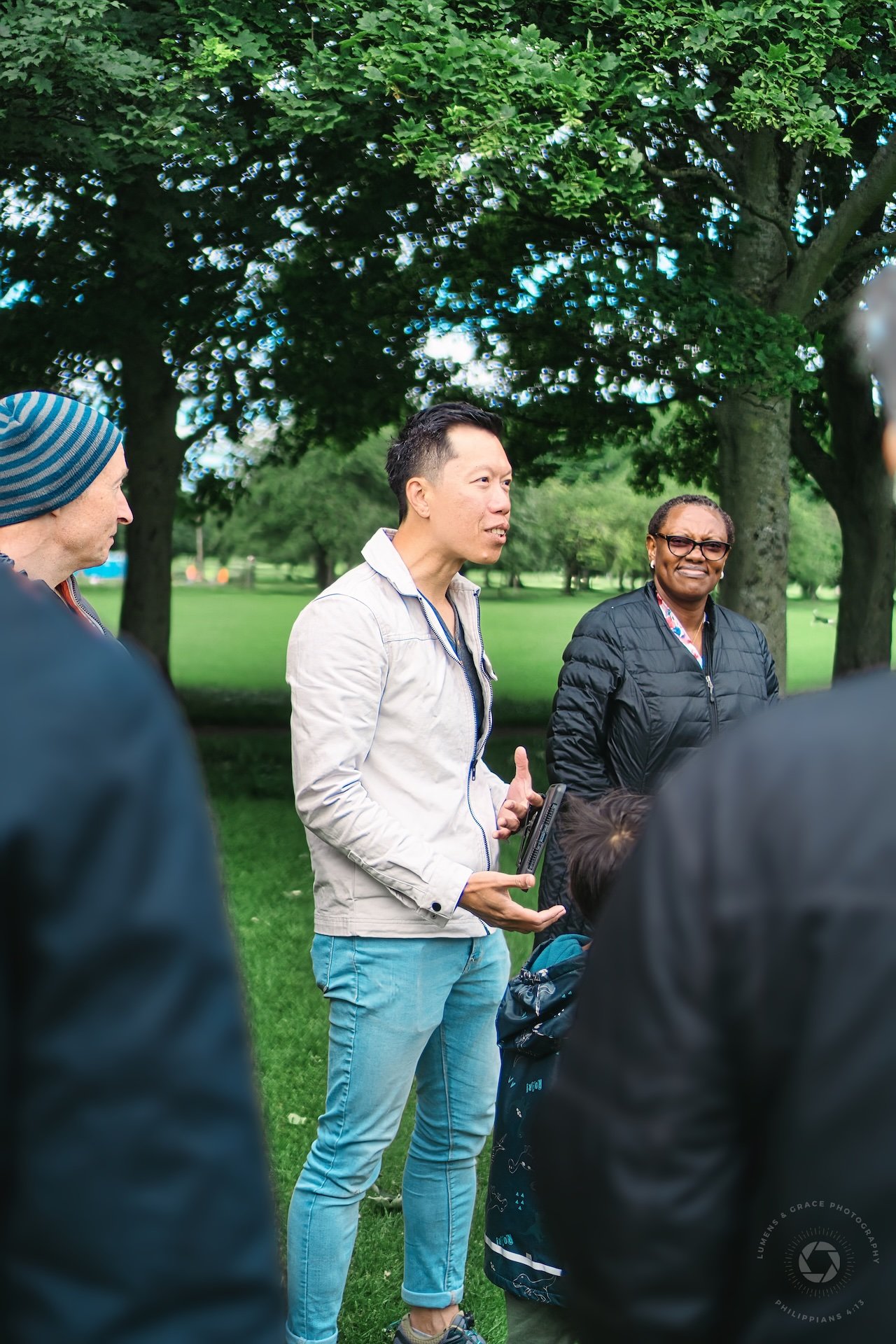 A group of people standing and talking in a park with trees and grass in the background.