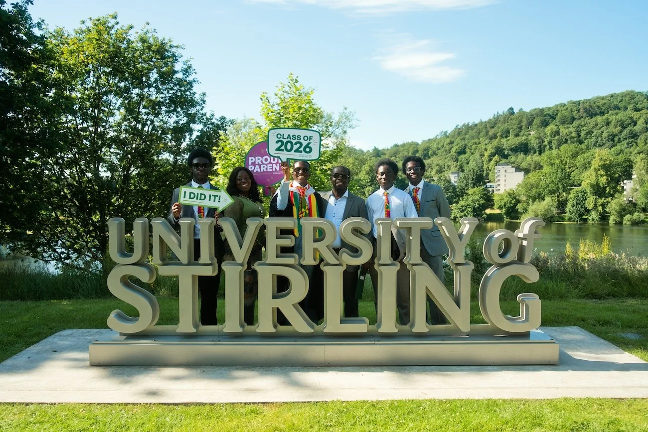 Group of seven young adults in graduation caps and gowns standing behind a large sign that reads 'University of Stirling' outdoors near a lake with trees and hills in the background.