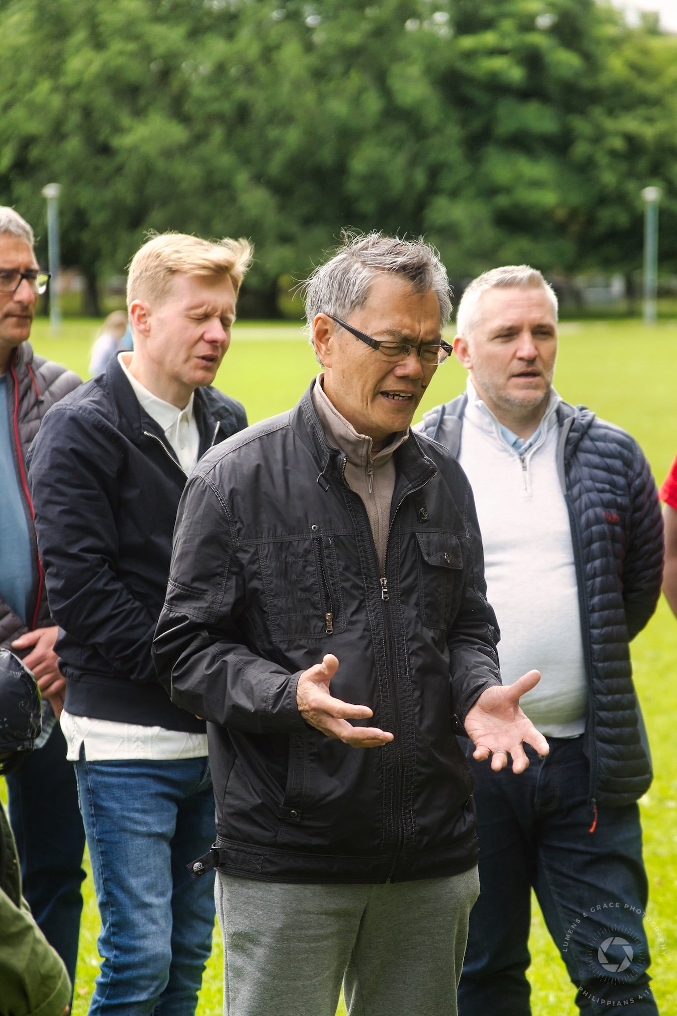 Group of men outdoors in a park, with trees in the background, engaged in a conversation or activity.