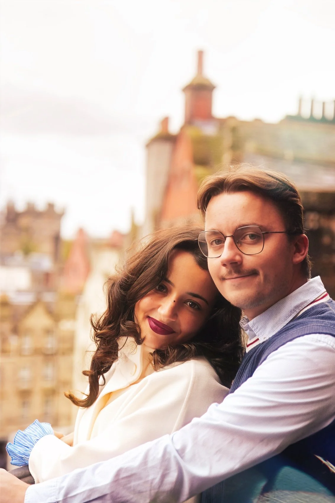 A young couple close together outdoors with city buildings and rooftops in the background. The woman has dark hair and lipstick, and the man has glasses and light skin.