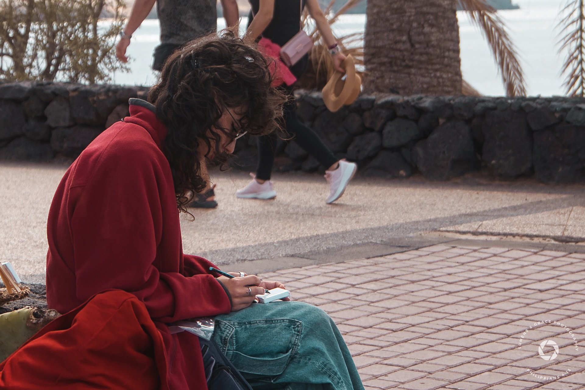 Person wearing a red coat, seated outdoors, writing or drawing in a notebook with casual pedestrians and palm trees in the background.