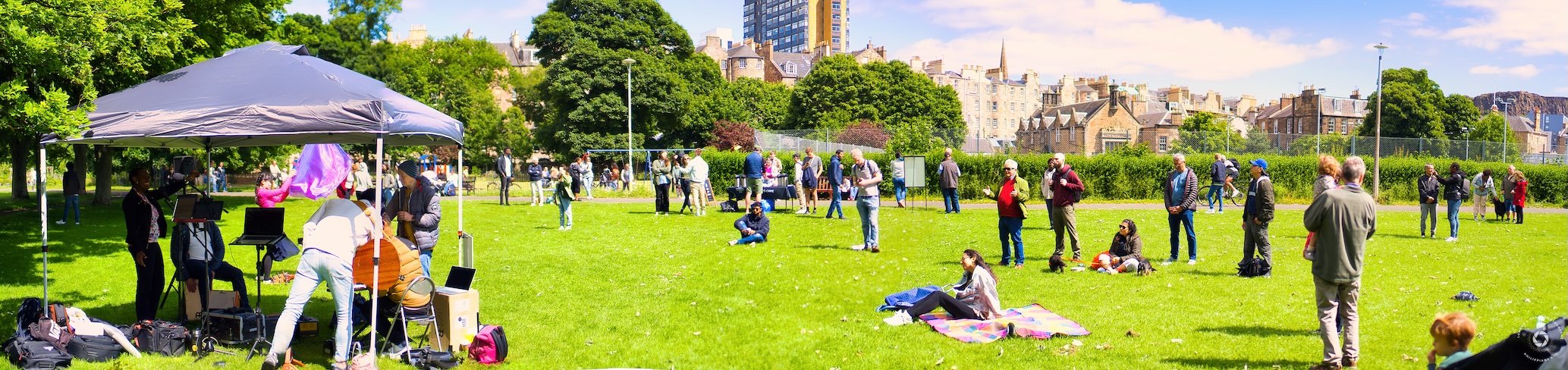 Outdoor gathering in a park with people under a canopy, using microphones and sound equipment. Others are standing or sitting on the grass, with trees and buildings in the background on a sunny day.
