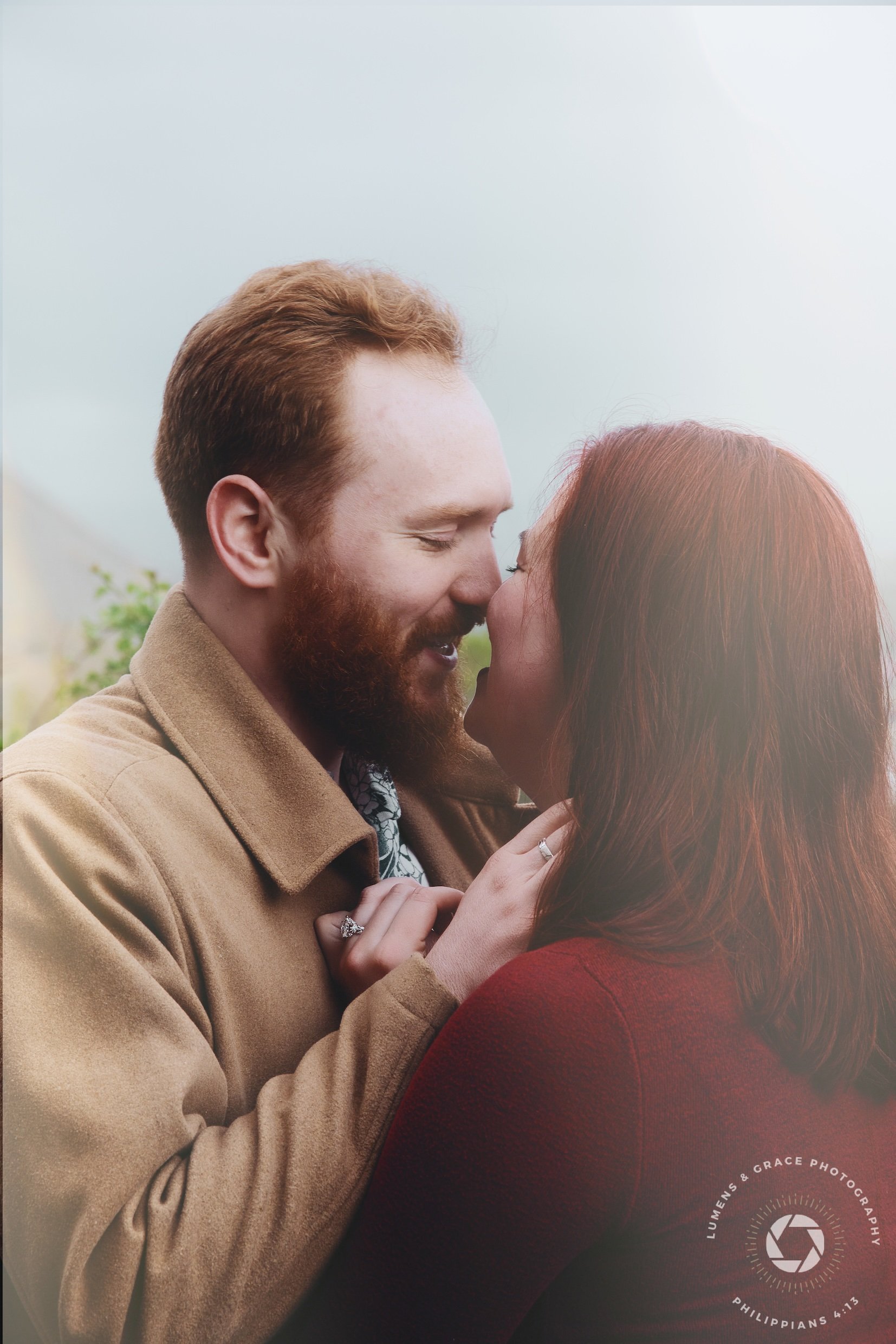 A couple embracing closely, smiling with eyes closed, outdoors with a blurred background. The man wears a tan coat and the woman wears a red top.