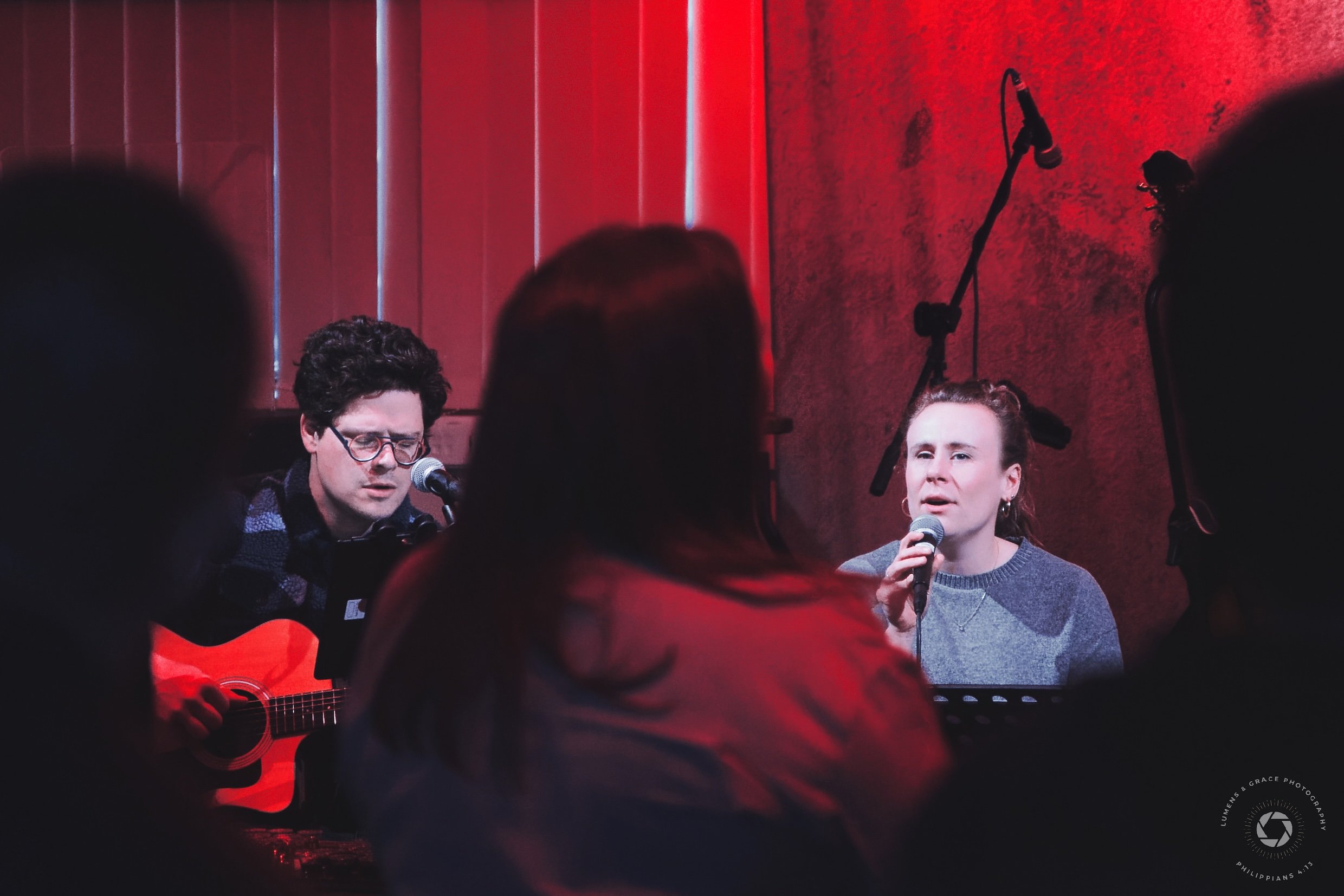 Two musicians performing on stage with red lighting, one playing guitar and singing, the other holding a microphone. Audience partially visible in foreground.