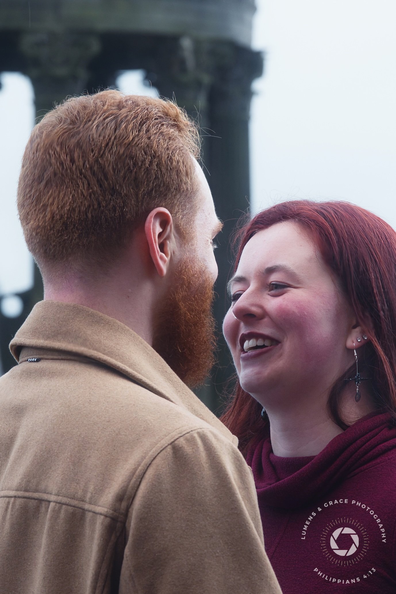 A close-up of a couple smiling at each other. The man has red hair and a beard, wearing a tan coat. The woman has red hair and is wearing earrings and a maroon top. Background includes out-of-focus stone pillars.