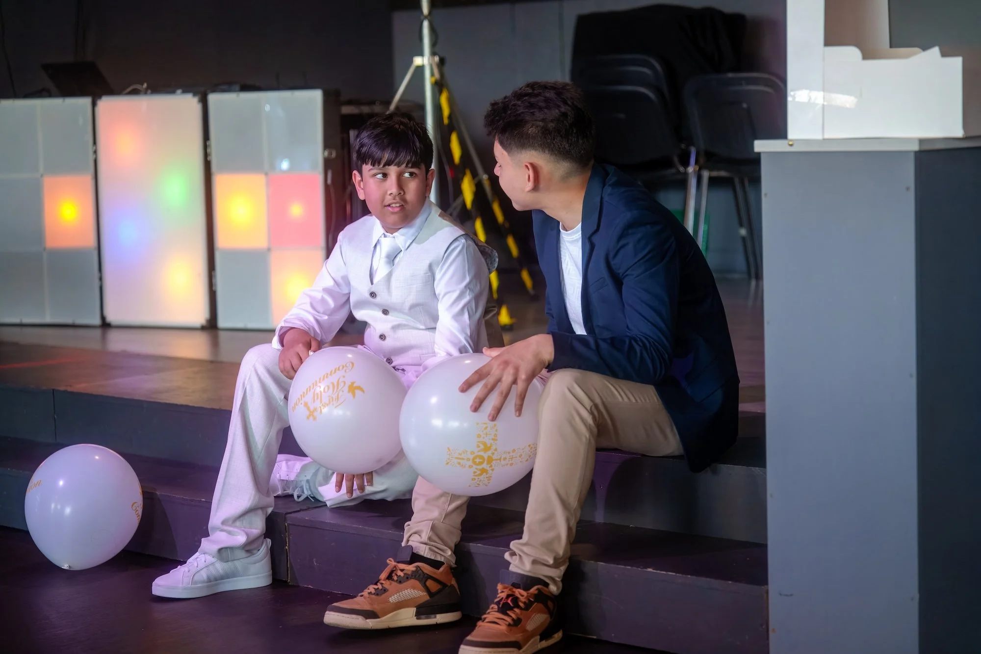A boy dressed in a white suit sitting on a stage holding white balloons while a man kneels beside him, both having a conversation. There are more balloons on the floor and colorful lighting in the background.