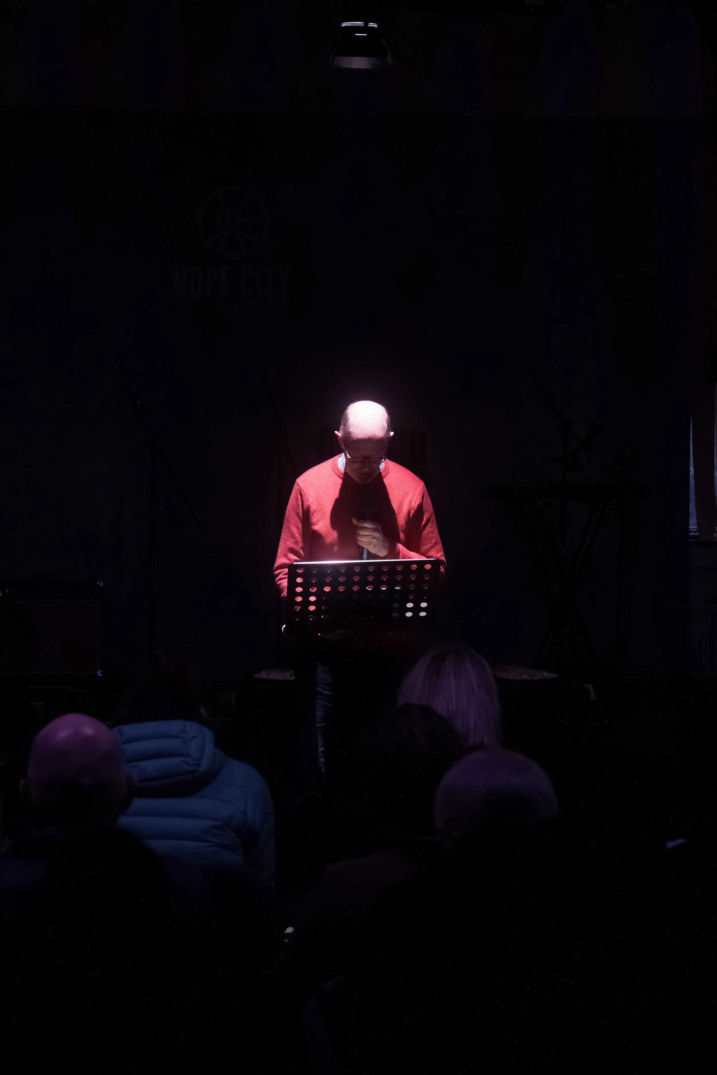 A man with glasses wearing a red sweater is standing on a dark stage, holding a microphone, with stage lights above him. The audience is visible in the foreground.