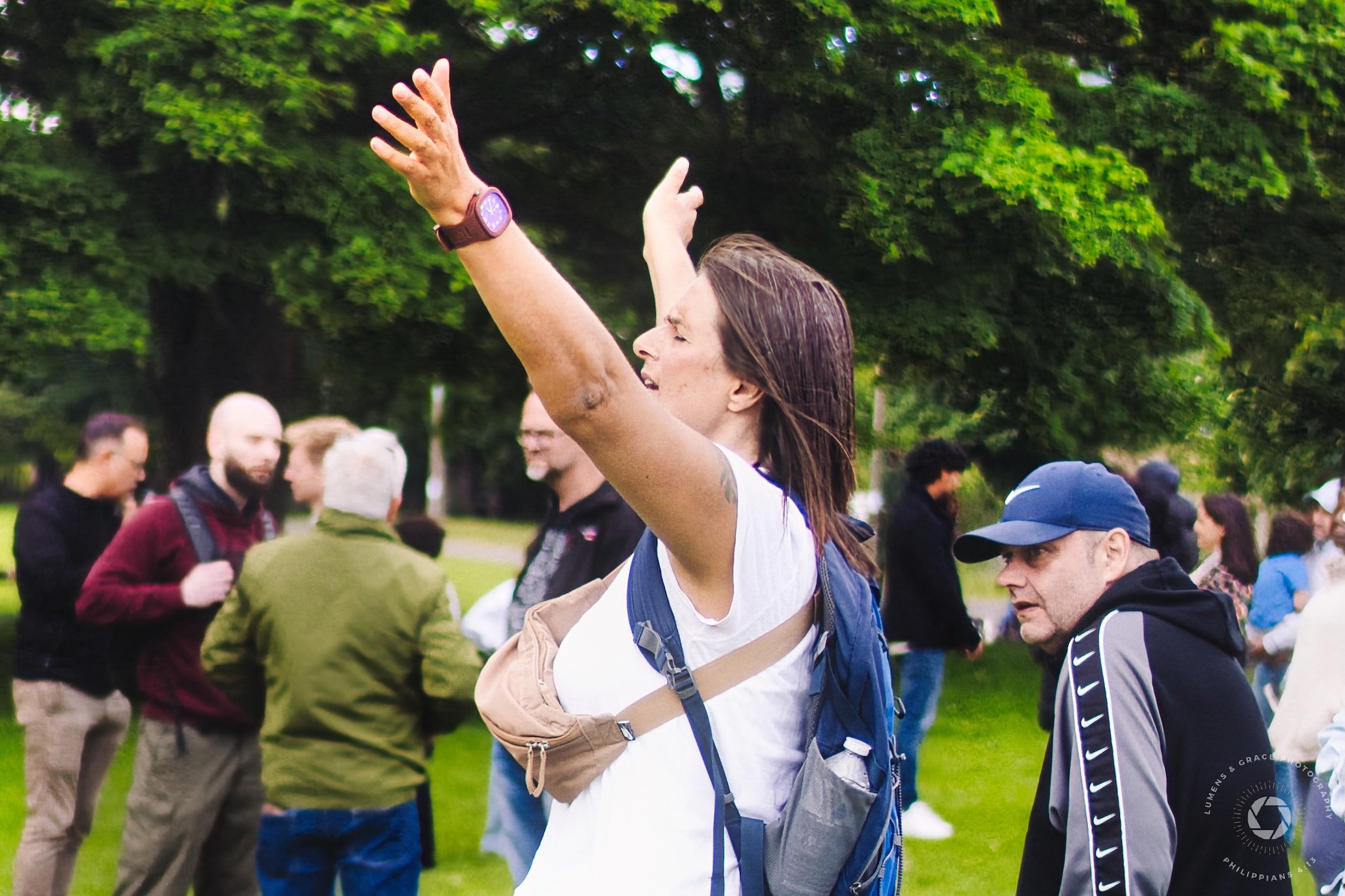 Person with arms raised in outdoor gathering, wearing a backpack and fanny pack, surrounded by people and trees.