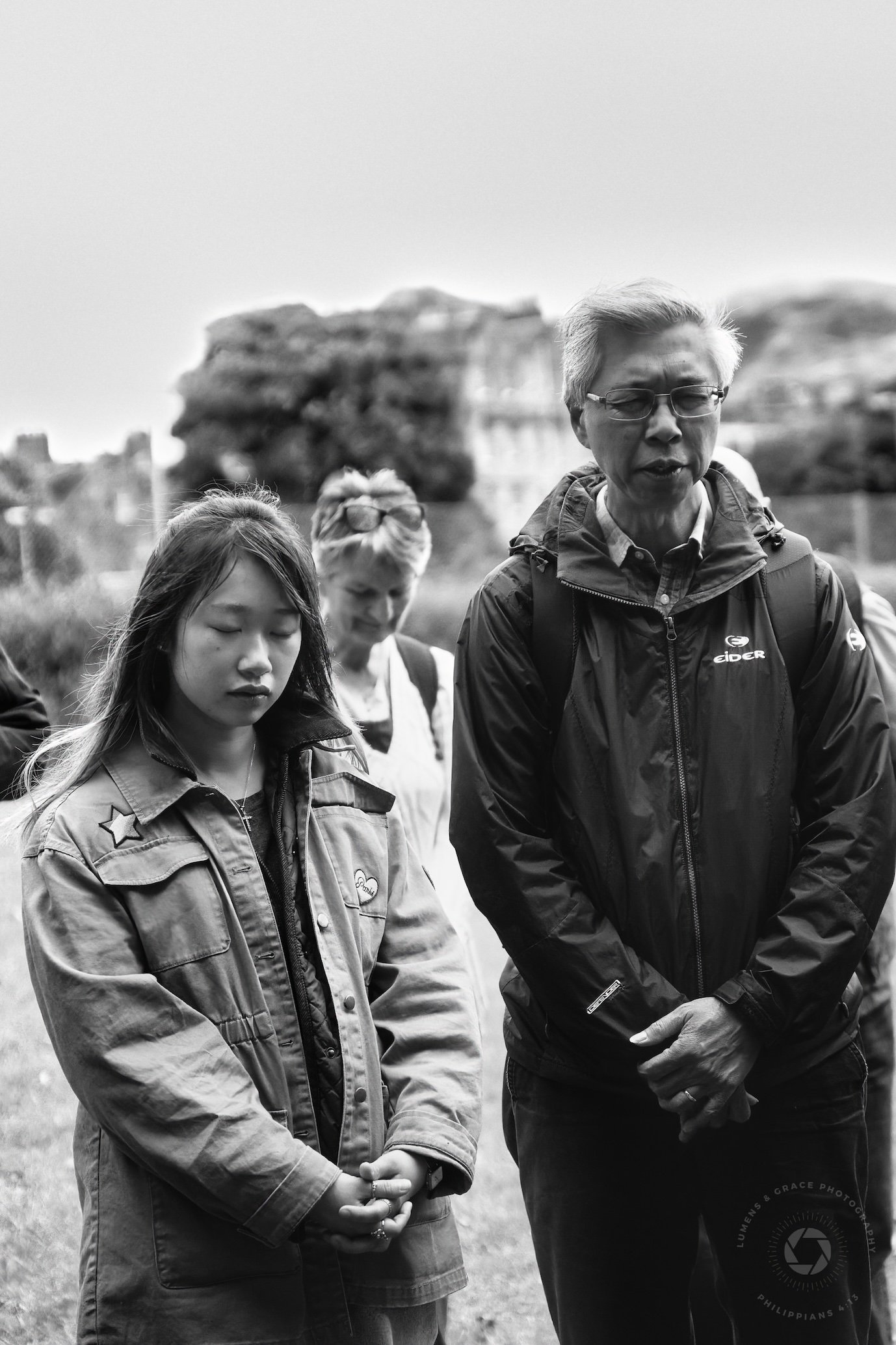 People standing outdoors, with some appearing to have their eyes closed, possibly in a contemplative or reflective mood.