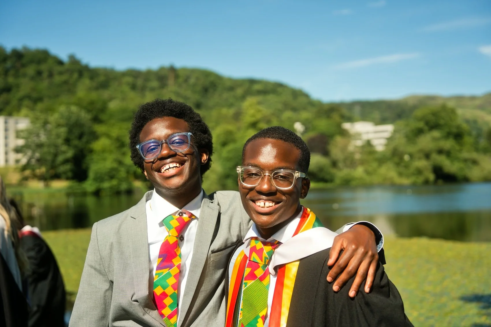 Two young men smiling and wearing colorful ties, celebrating outdoors near a body of water with green hills in the background.