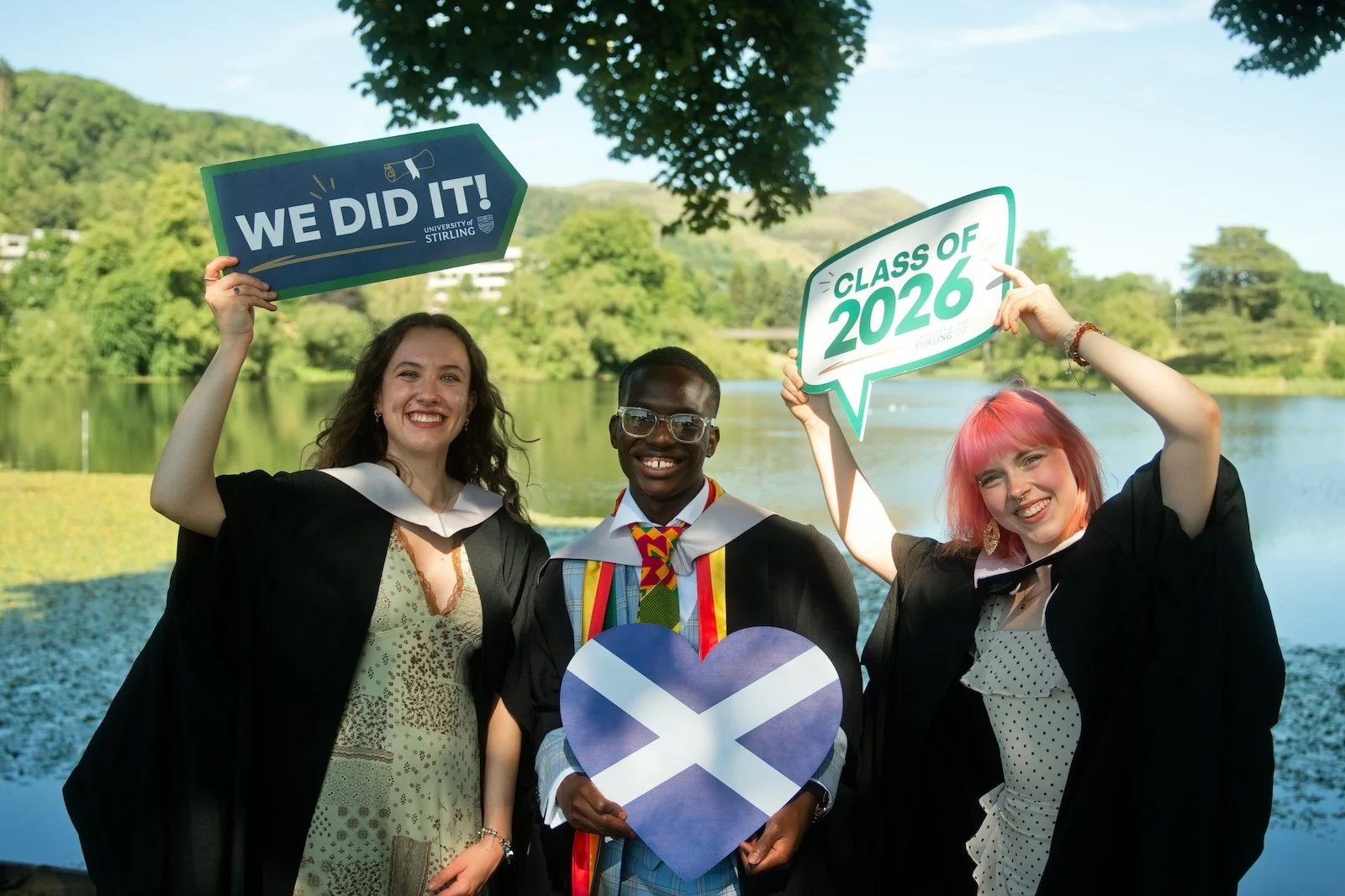 Three young adults in graduation gowns standing outdoors by a lake, smiling and holding signs that read 'We Did It!' and 'Class of 2026.' The graduate in the center is wearing a gown with rainbow-colored honor cords and holding a heart-shaped sign wi