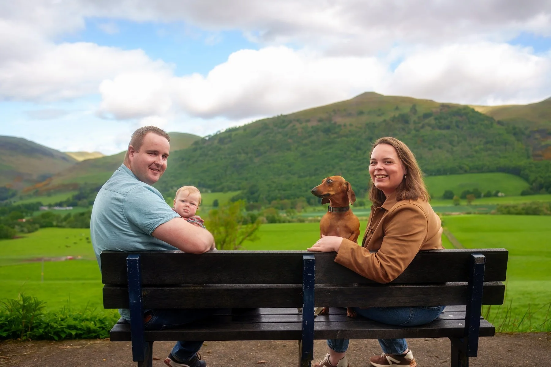 A family of four and their dog sitting on a park bench outdoors with green rolling hills and mountains in the background; the father holding a baby, the mother smiling, and the dog sitting between them.