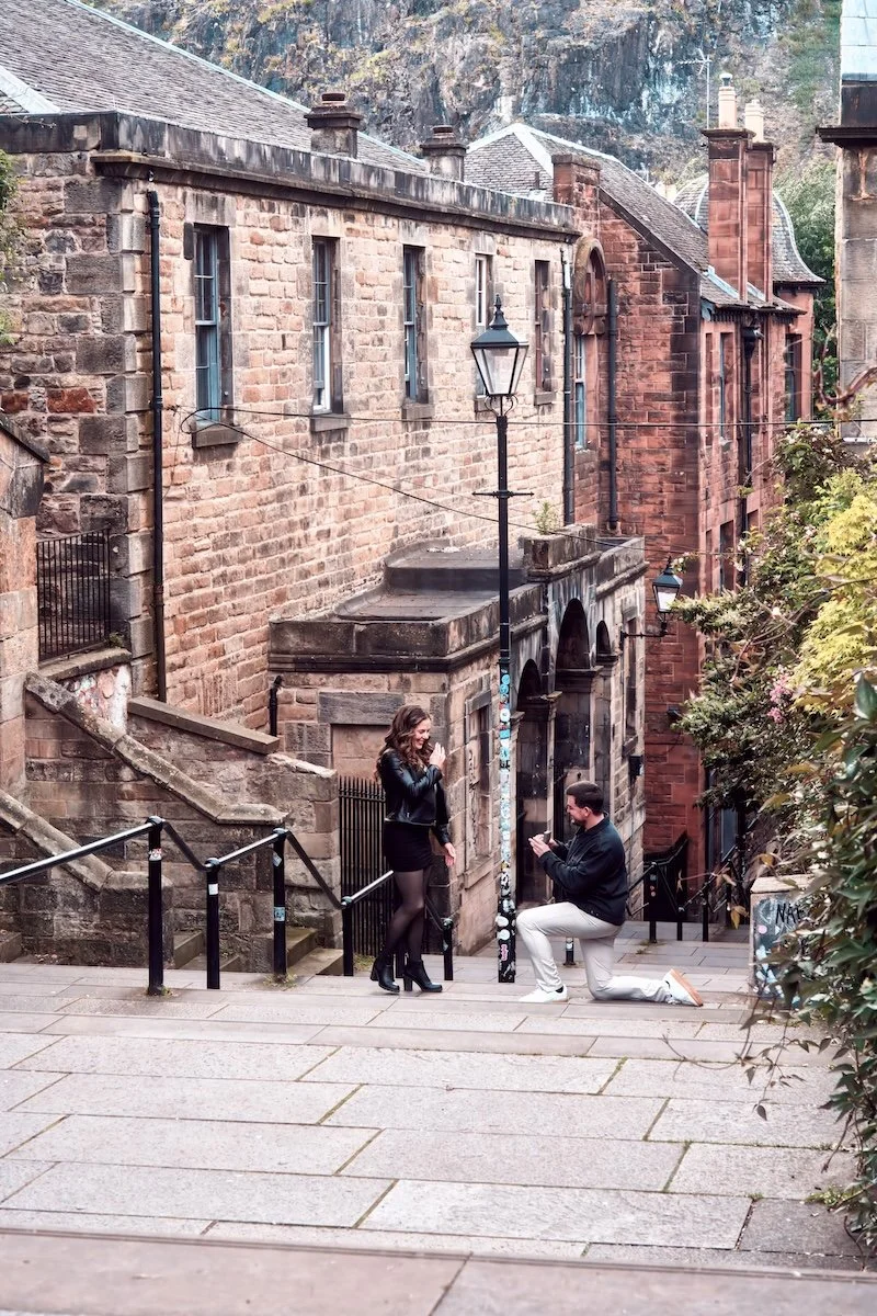 A man proposing marriage to a woman in a city street, with historic brick buildings and stairs nearby.