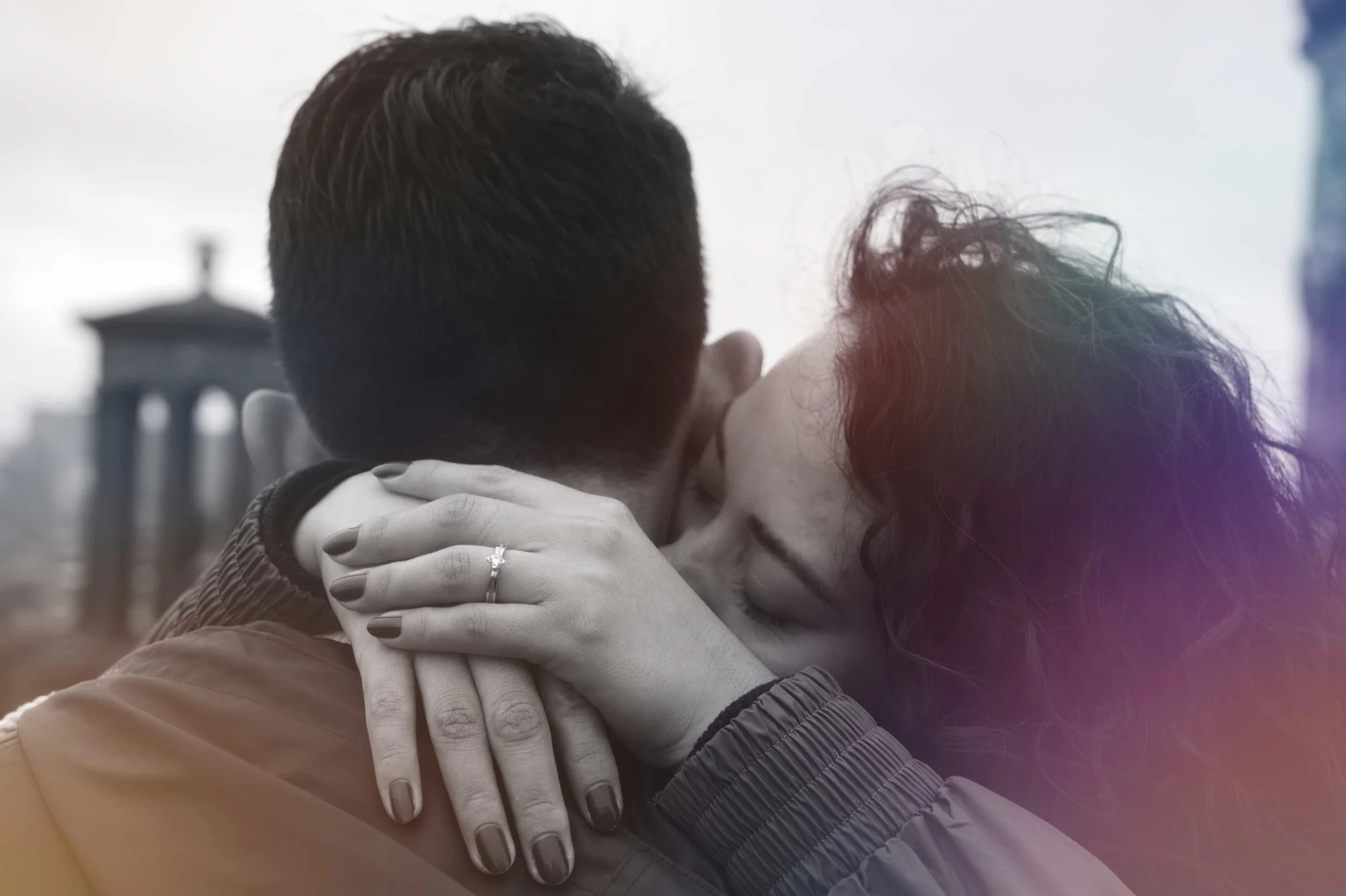 A couple sharing a romantic embrace, holding each other closely with their heads touching, outdoors with a blurred cityscape in the background.