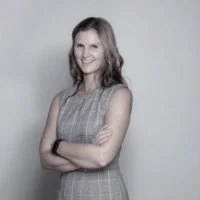 A woman with long hair smiling and crossing her arms, standing against a plain light gray background.