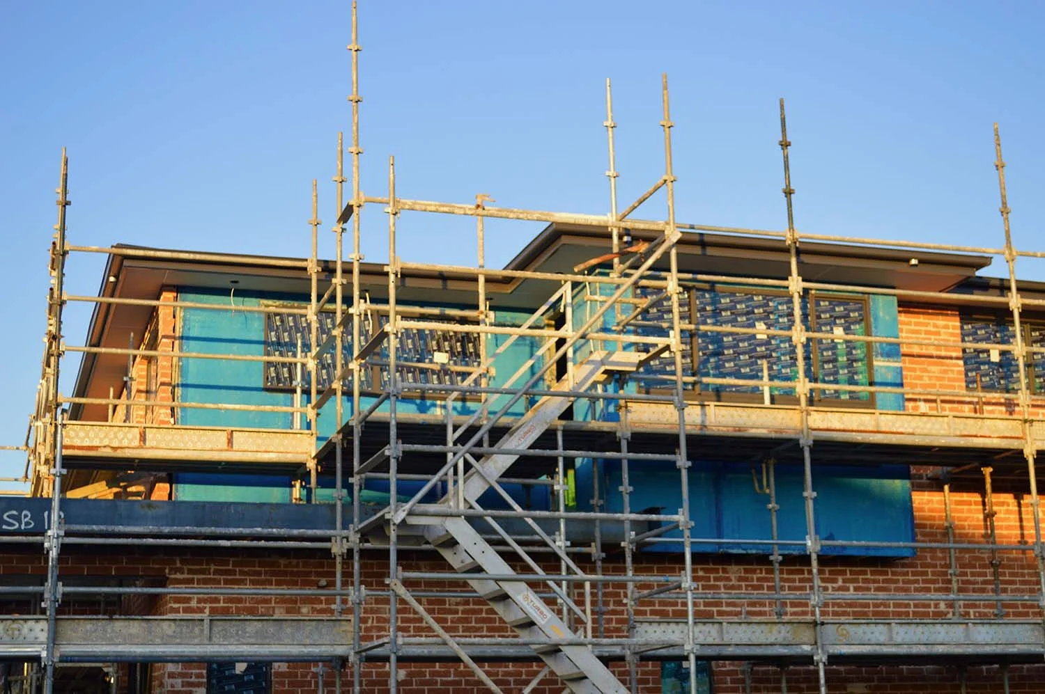 Funding for a construction site with scaffolding around a brick house under renovation, with visible blue insulation materials and a clear blue sky.