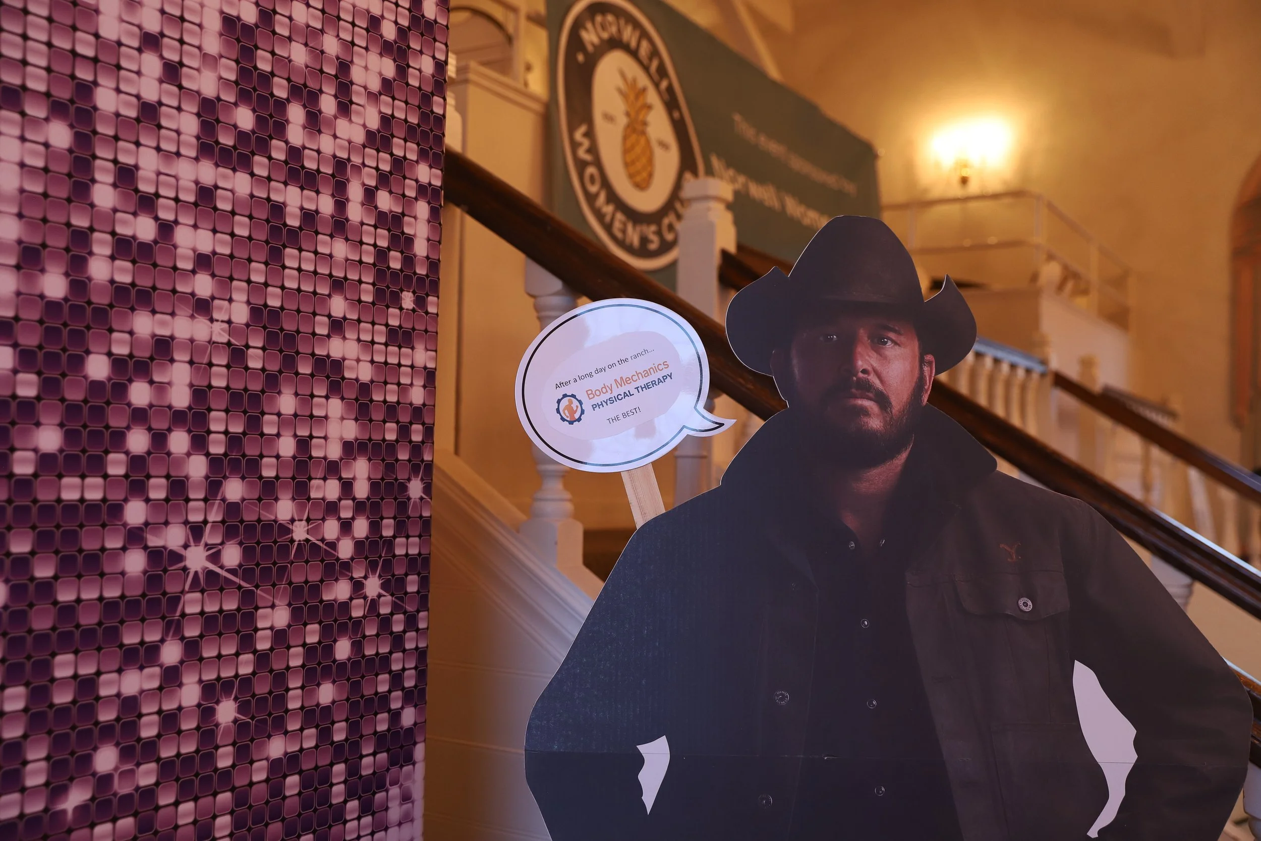 A cardboard cutout of a man wearing a black cowboy hat and black jacket, standing indoors near a staircase. There is a speech bubble indicating 'After a long day on the ranch... Body Mechanics Physical Therapy THE BEST!'. The background is purple met