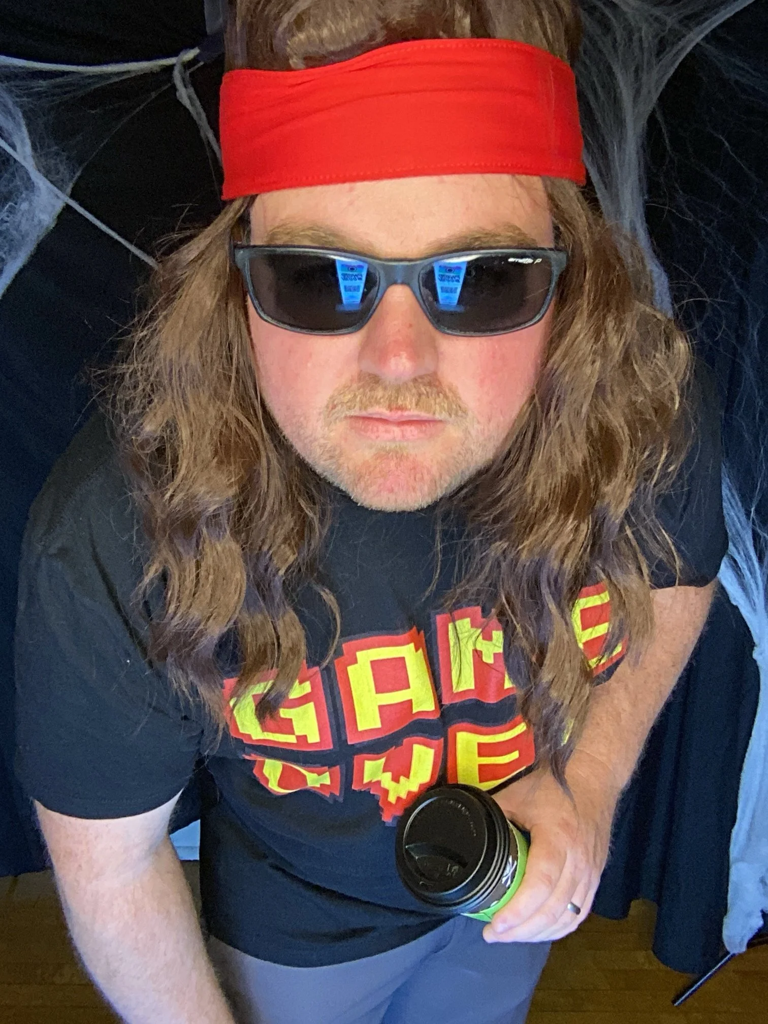 A man with long wavy hair wearing sunglasses, a red headband, a black T-shirt with colorful text, holding a travel mug and looking up toward the camera.