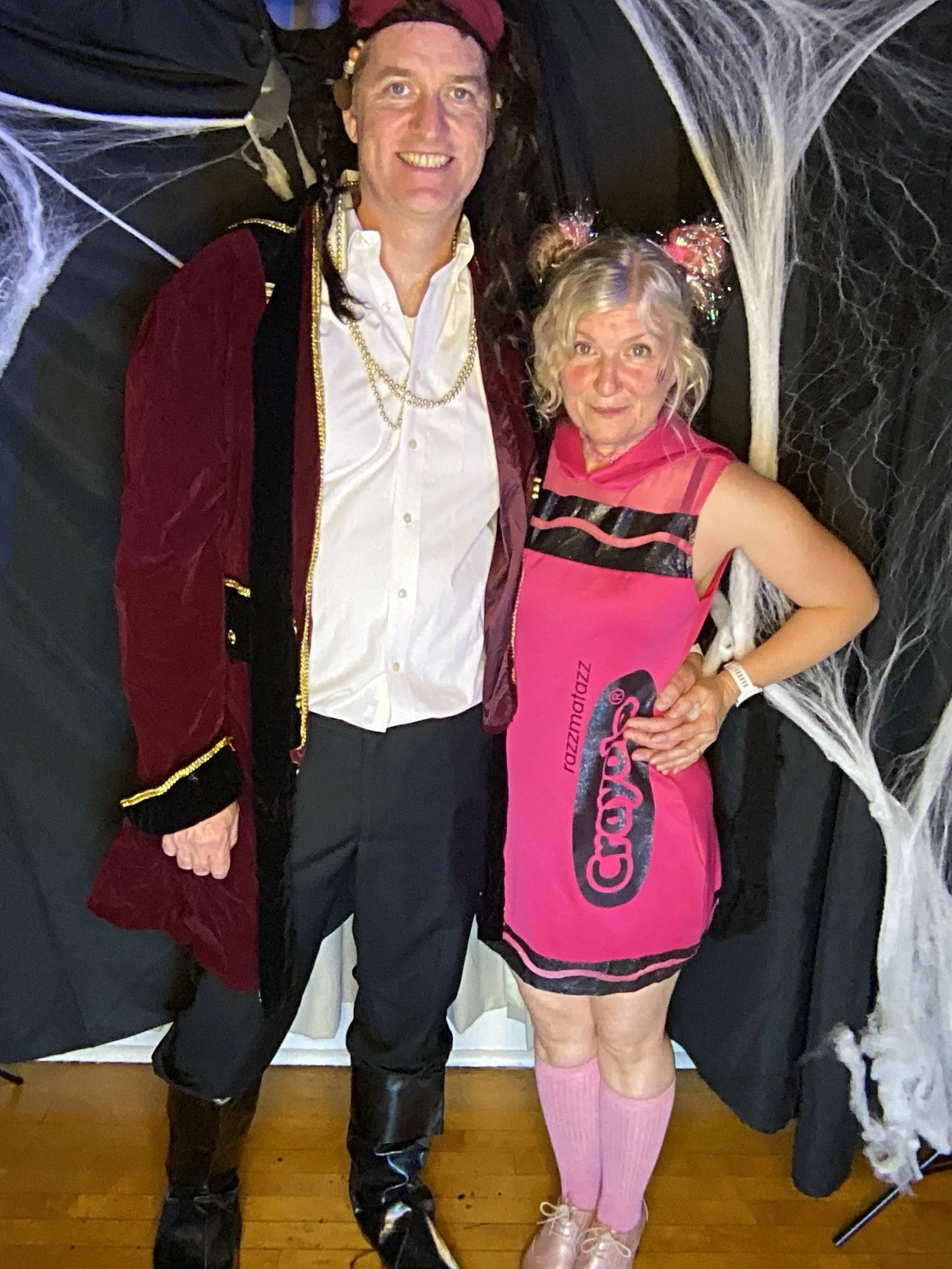 Two people dressed in costumes standing in front of a Halloween-themed backdrop with fake spider webs. The person on the left is wearing a pirate costume with a velvet jacket, white shirt, and black boots. The person on the right is wearing a pink dr