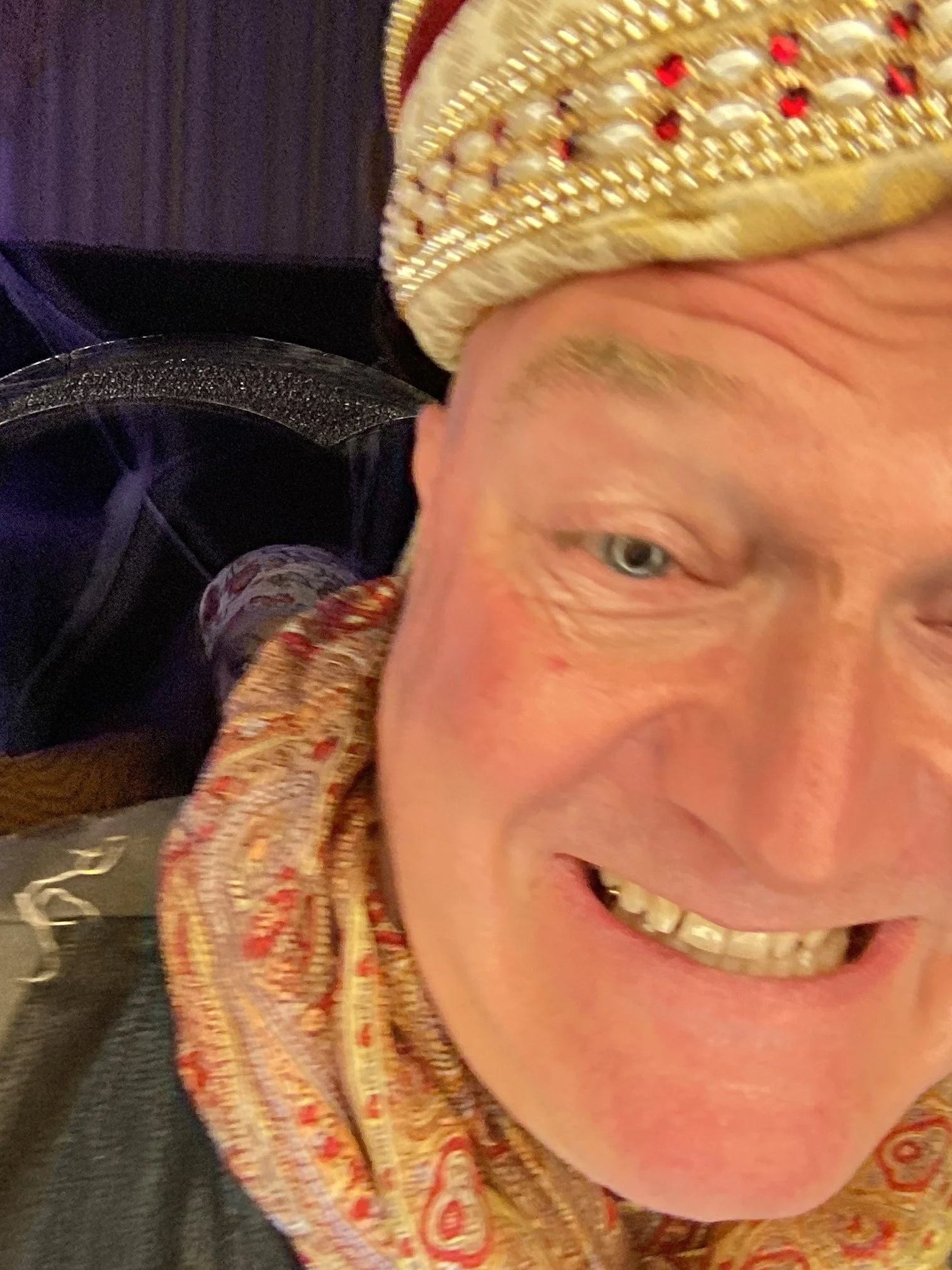 A close-up of a smiling man wearing a gold, pearl, and red-embellished crown, with a paisley scarf around his neck.