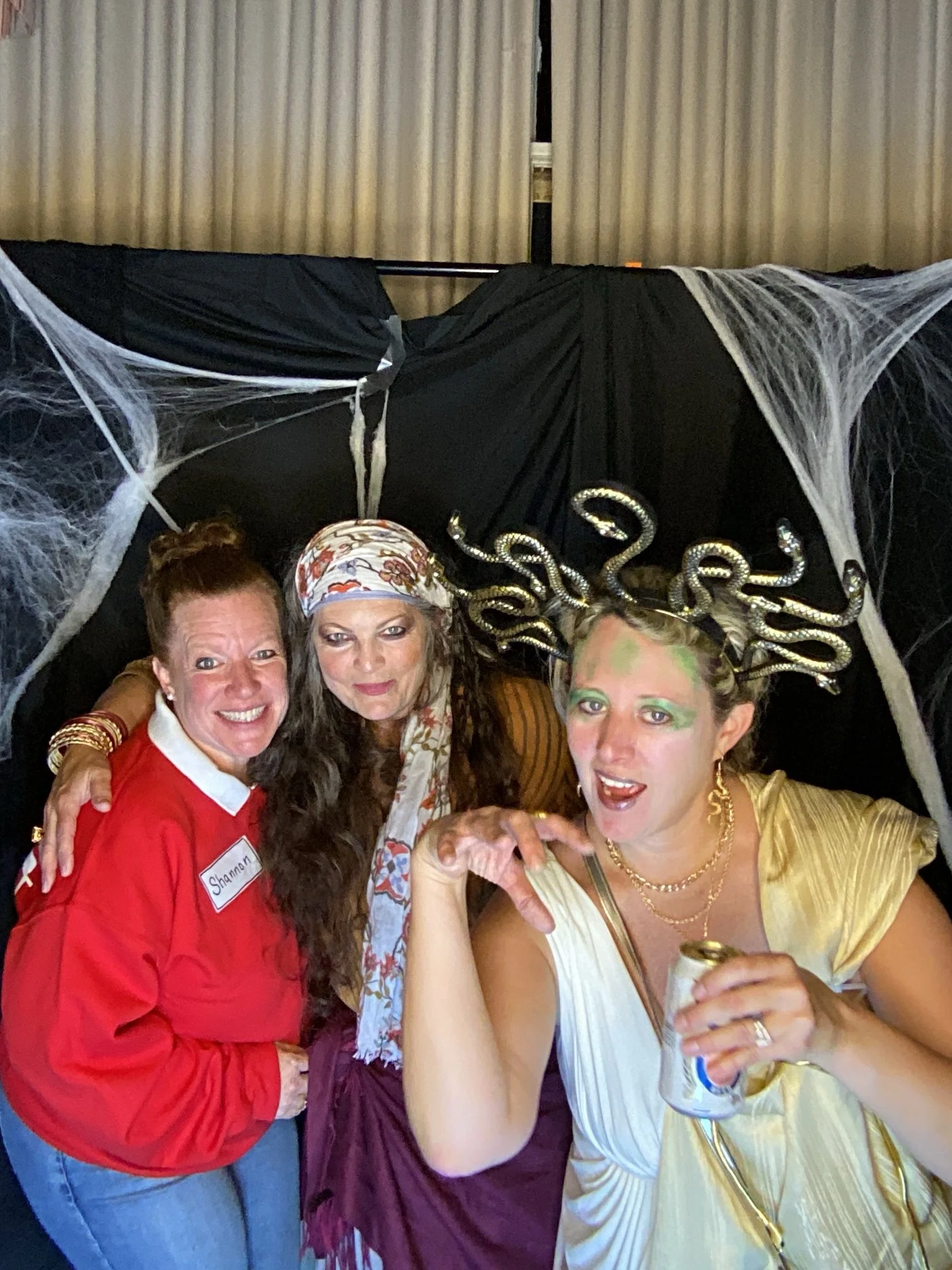 Three women dressed in costumes pose for a photo in front of black fabric decorated with spider webs. The woman on the left wears a red shirt and has a name tag that reads "Shannon." The woman in the middle has long dark hair, a colorful headscarf, a