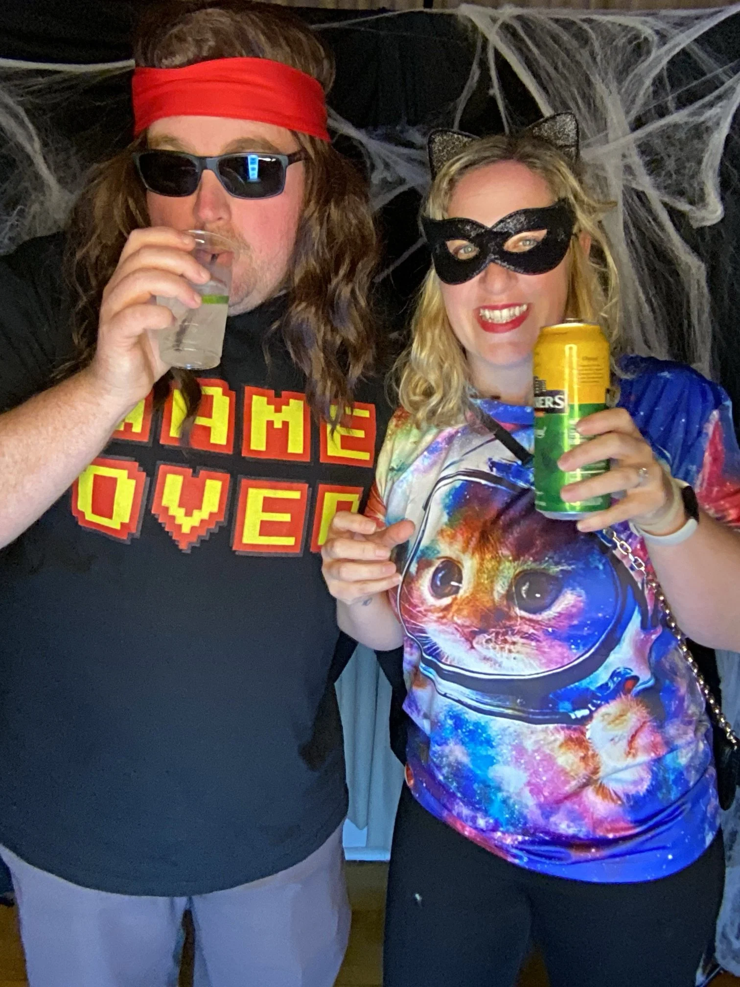 Two people dressed in costumes – the man wearing sunglasses, a red headband, and a shirt that says 'GAME OVER', and the woman wearing a black cat mask, cat ears, and a colorful space-themed shirt with a cat image – holding drinks and posing for a pho