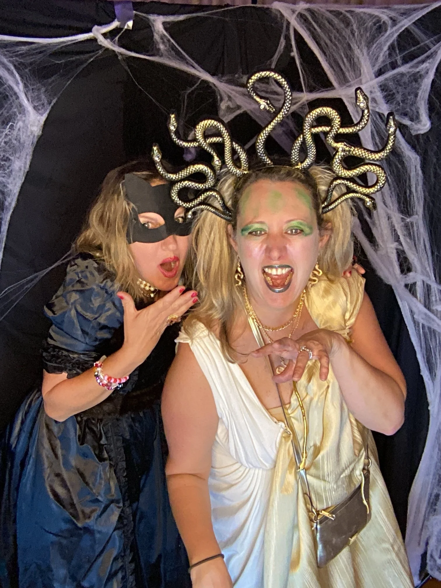 Two women in costumes posing in front of a spiderweb backdrop. The woman on the right is wearing a shiny gold dress, jewelry, and a headpiece with snake-shaped decorations. The woman on the left is dressed as a thief or ninja with a black mask and da