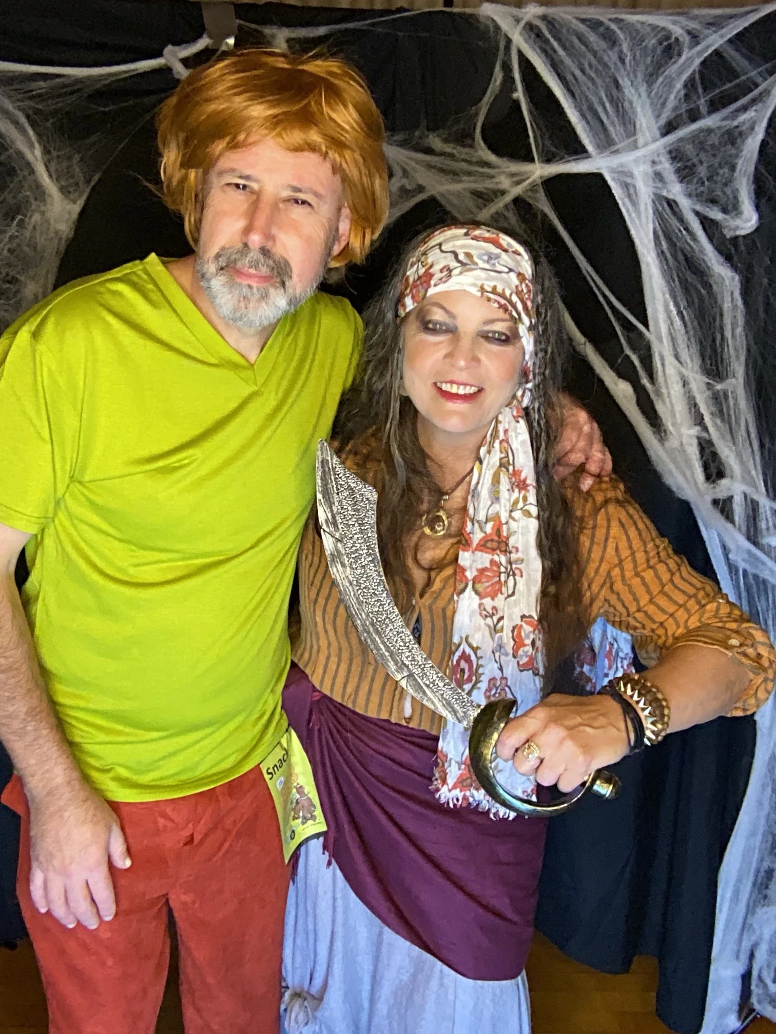 Two people in costumes, one man dressed as a hiker with a red beard and wig, the other woman in pirate costume holding a sword, standing in front of a backdrop decorated with fake cobwebs for Halloween.