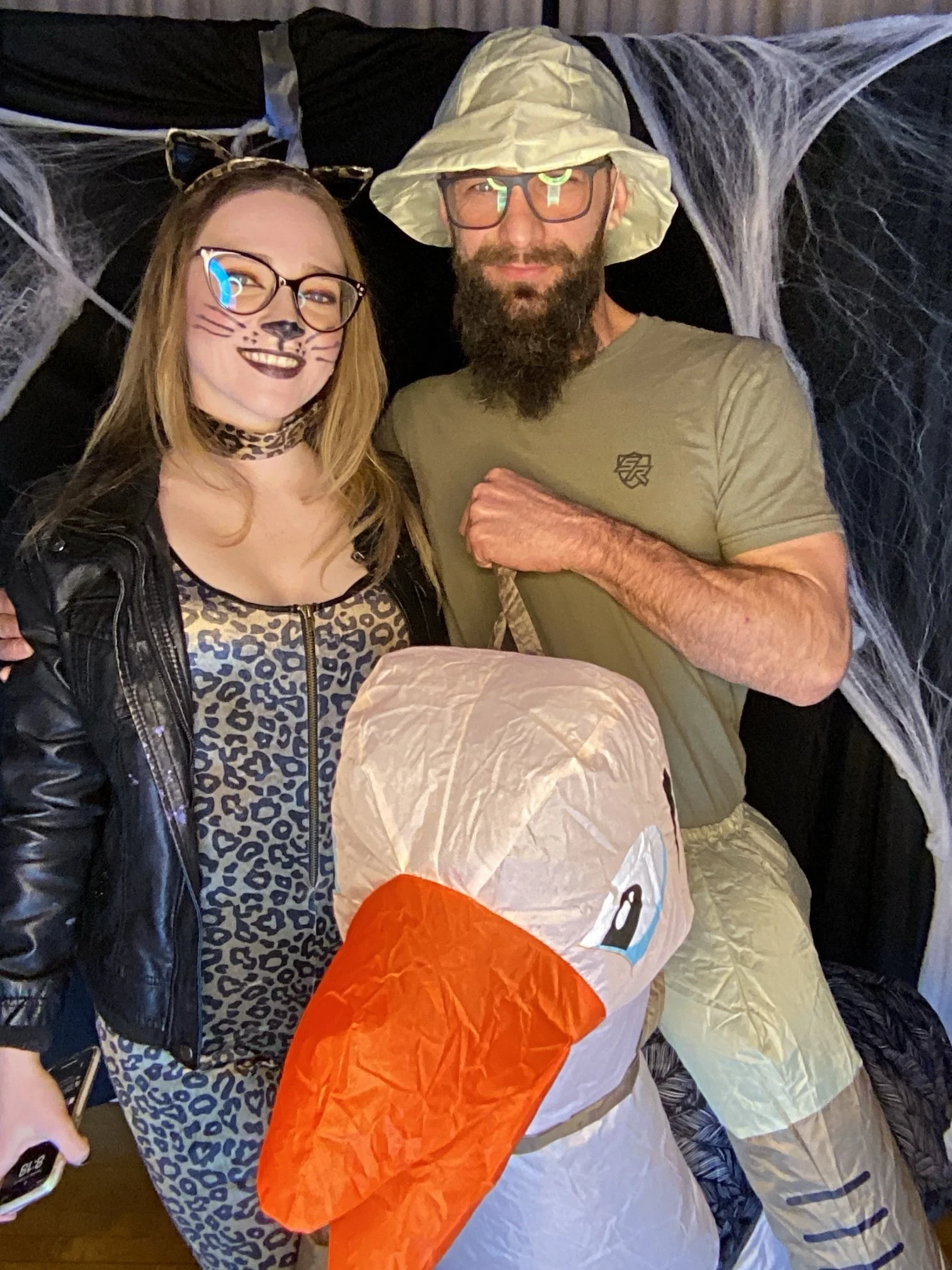 Two people dressed in costumes standing behind a large inflatable duck with a white body and orange beak. The woman on the left has face paint resembling a cat, with a black nose, whiskers, and a leopard print choker. The man on the right is wearing