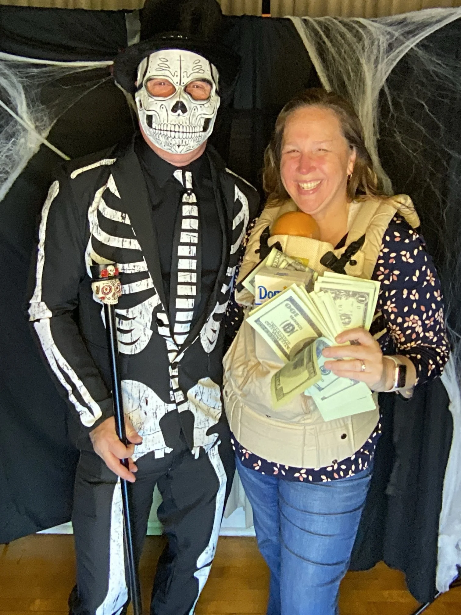 A man dressed in a skeleton costume with a skull mask and a woman smiling, holding dollar bills, in front of Halloween decorations.