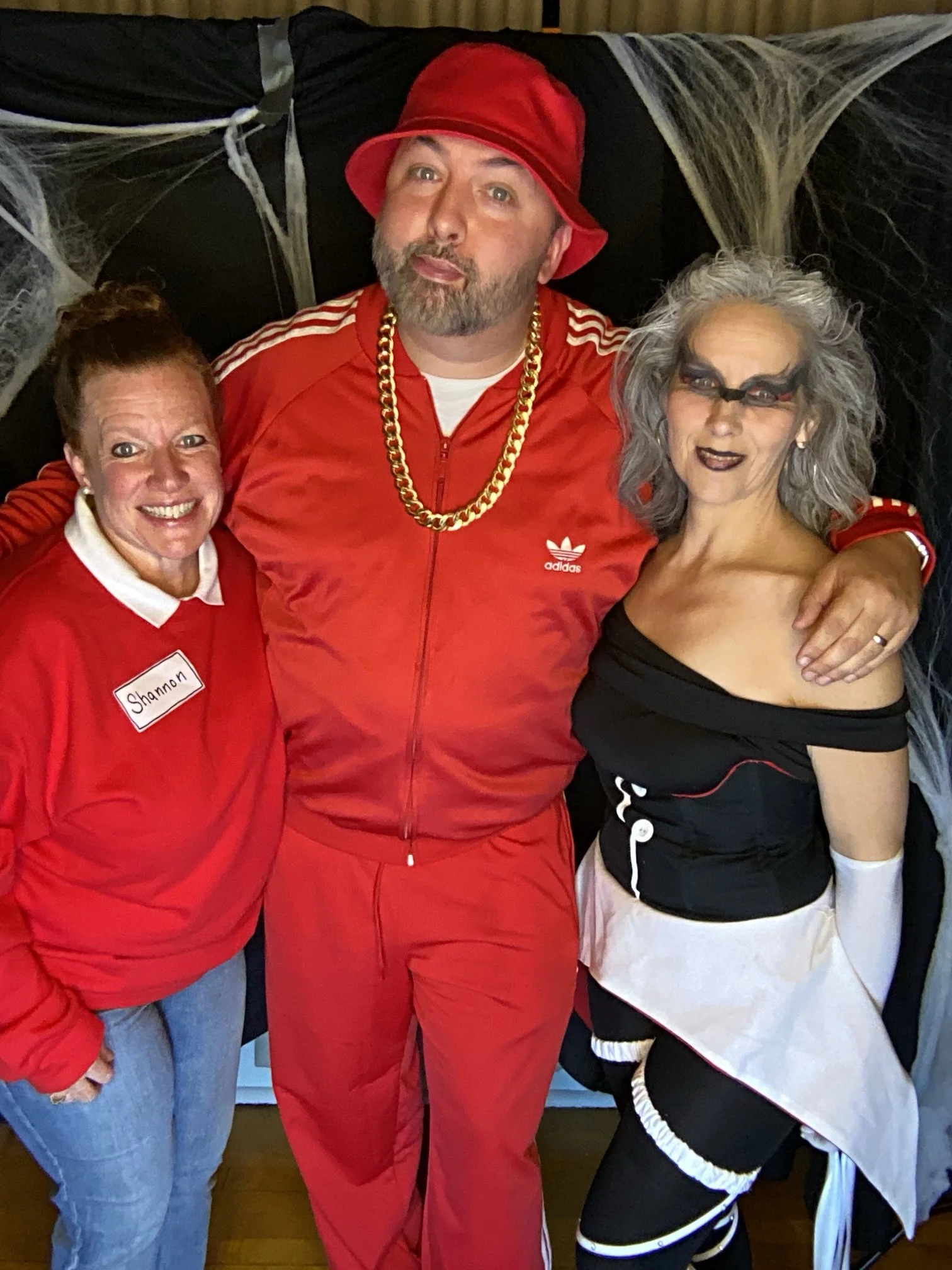 Three people dressed in costumes posing together for a photo. The woman on the left is wearing a red jacket with a name tag that reads 'Shannon' and has reddish hair. The man in the middle is dressed in a red tracksuit, a red bucket hat, a gold chain