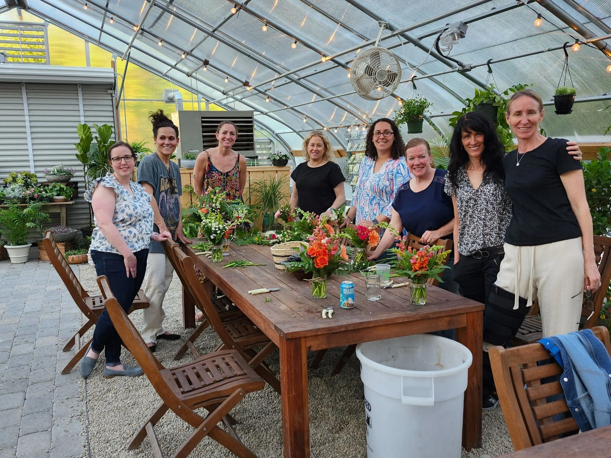 A group of eight women standing around a table with floral arrangements in a greenhouse, with various potted plants and flowers in the background.