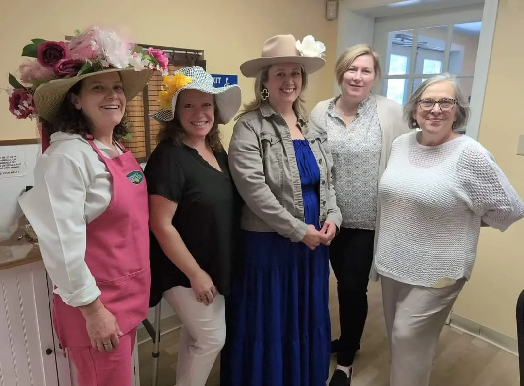 Group of five women standing indoors, smiling, some wearing large, decorative hats with flowers and ribbons, dressed in colorful and casual clothing.