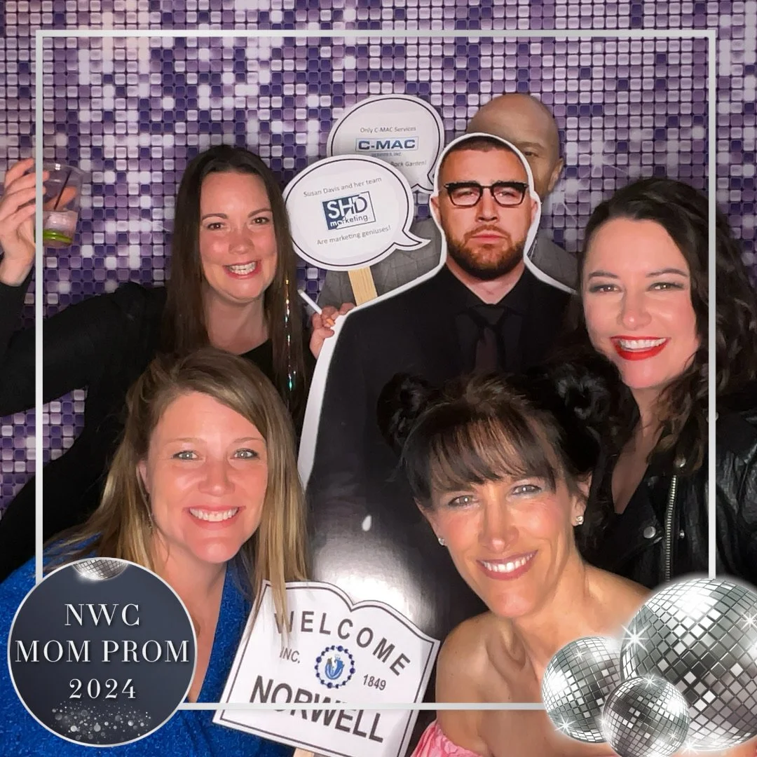 Group of women and one man posing at a prom event, holding signs and memorabilia, with a purple mosaic backdrop, themed for 'NWC MOM PROM 2024', decorated with disco balls.