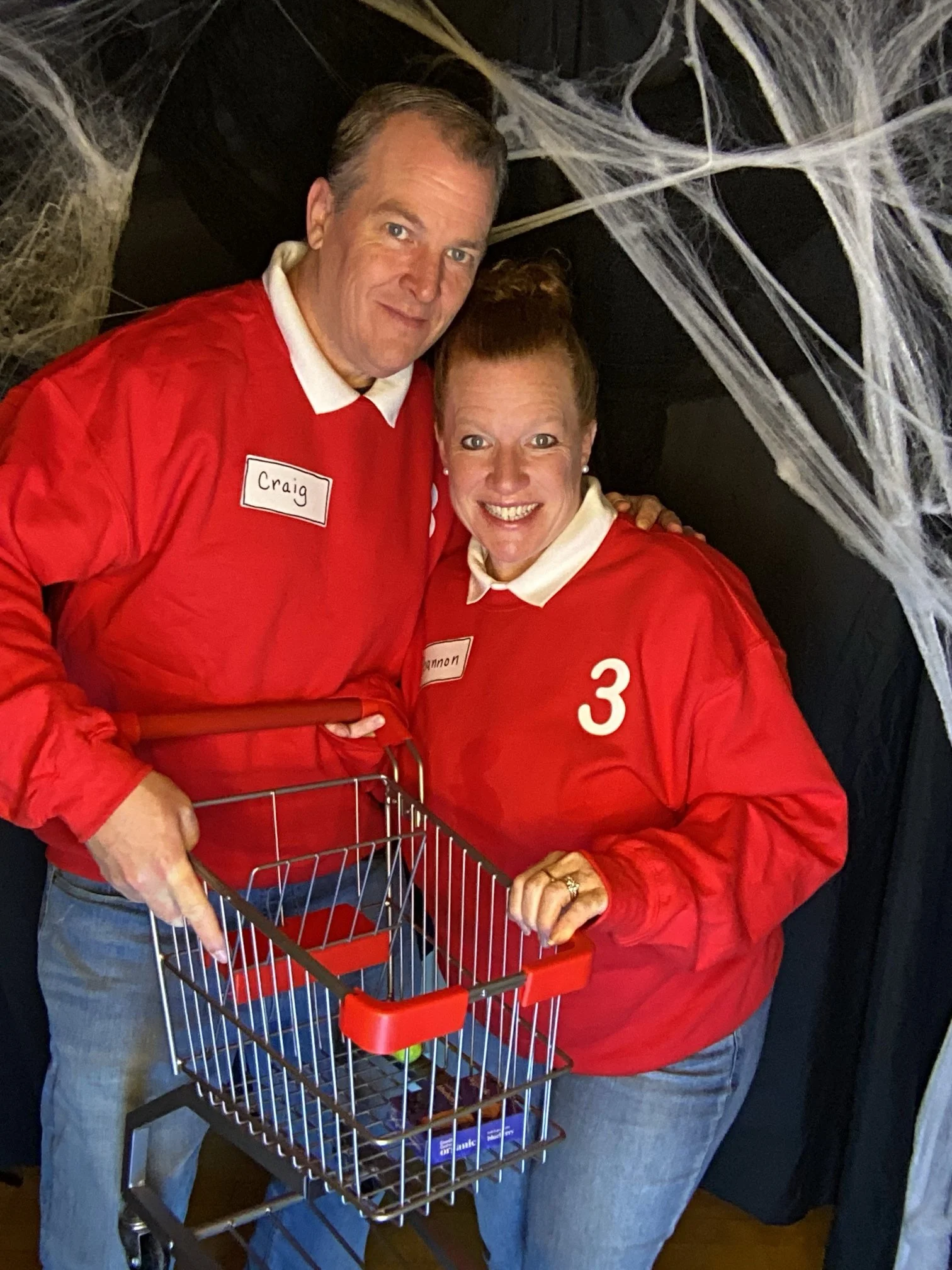 A man and woman dressed in red and white sports jerseys with name tags standing together, holding a small shopping cart with a Halloween-themed background of fake spider webs behind them.