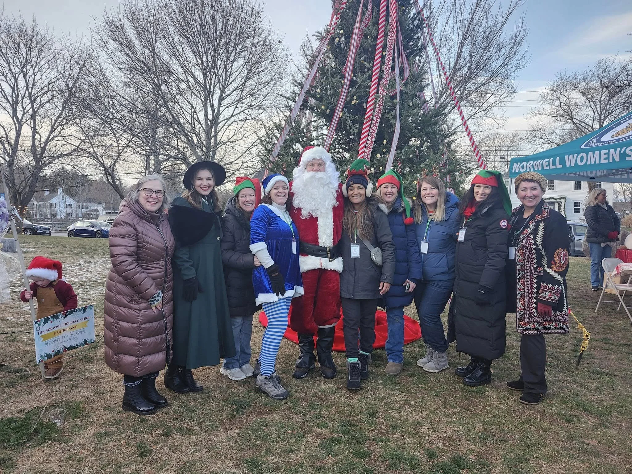 A group of women and Santa Claus standing in front of a decorated Christmas tree outdoors, with some wearing festive elf hats, a woman dressed as a holiday witch, and a child in a Santa hat and coat holding a sign. The scene is set in a park with tre