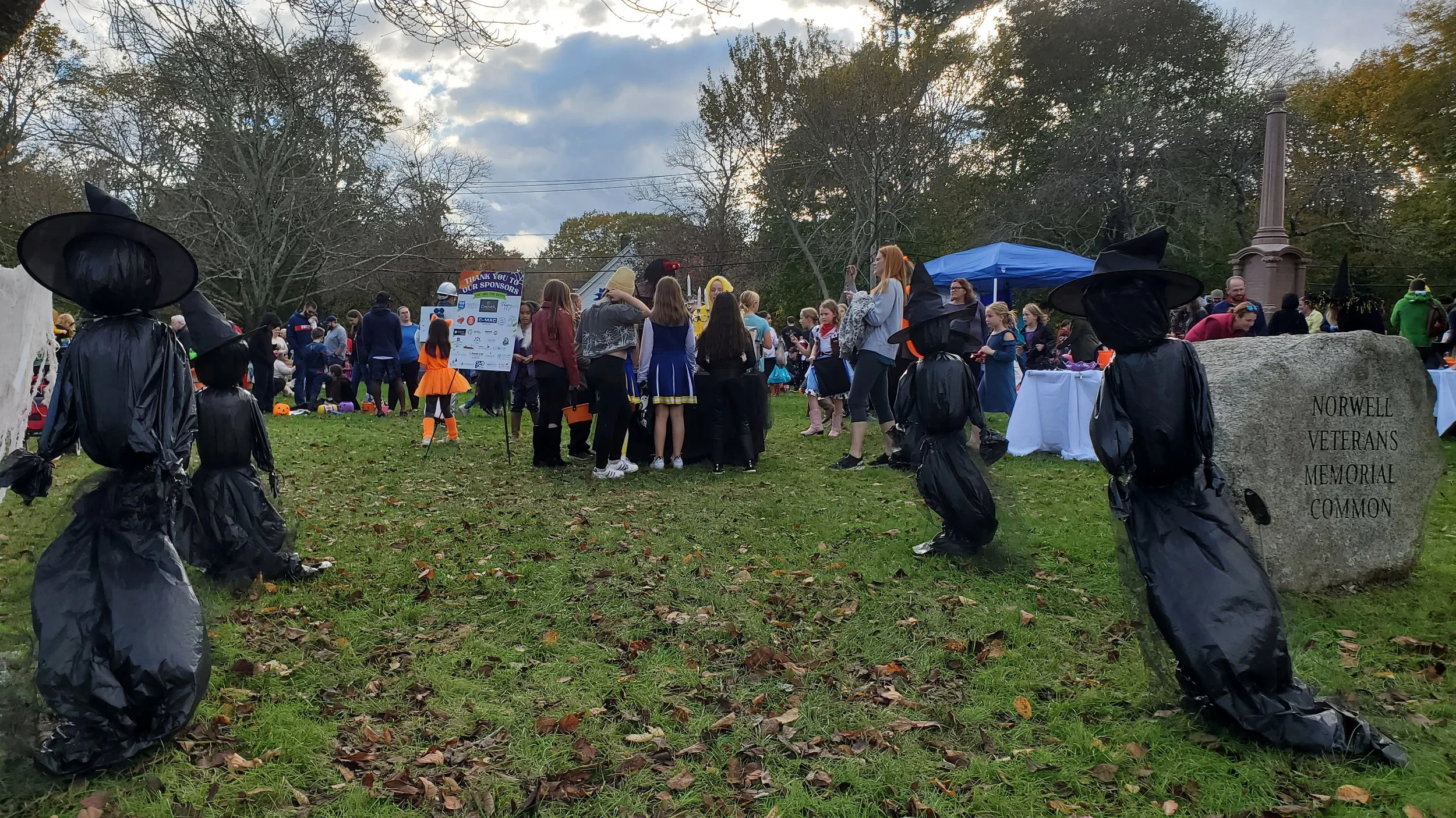 People gathered at a park during a Halloween event, with costumes and Halloween decorations, near the Norwell Veterans Memorial, with some scarecrow-like figures made of black plastic bags and a sign recognizing sponsors.