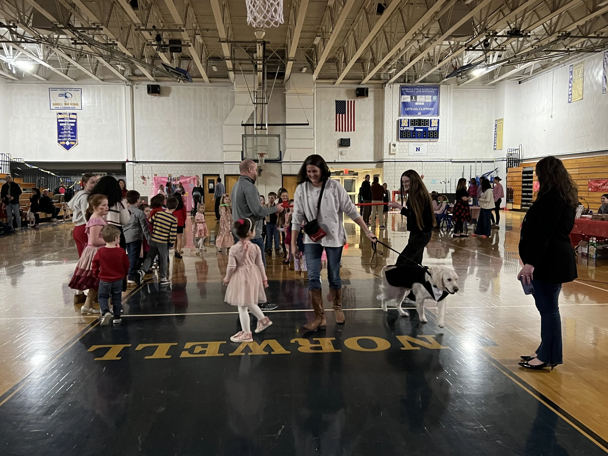 People and children in a gymnasium, some lined up for an event, with a woman walking a dog in the center.