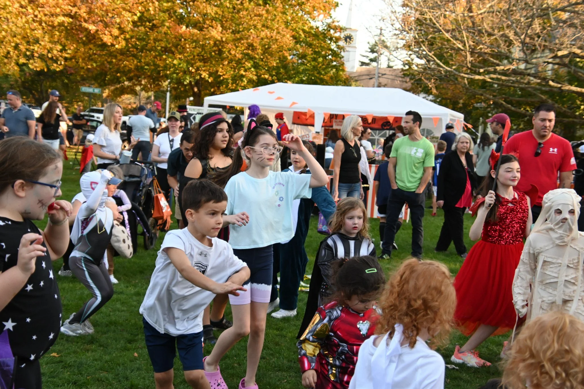 Children and adults at a Halloween outdoor event with some kids in costumes, including a girl with cat ears and face paint, a girl in a red dress with a headband and a girl in a mummy costume, with a white tent in the background and fall trees.