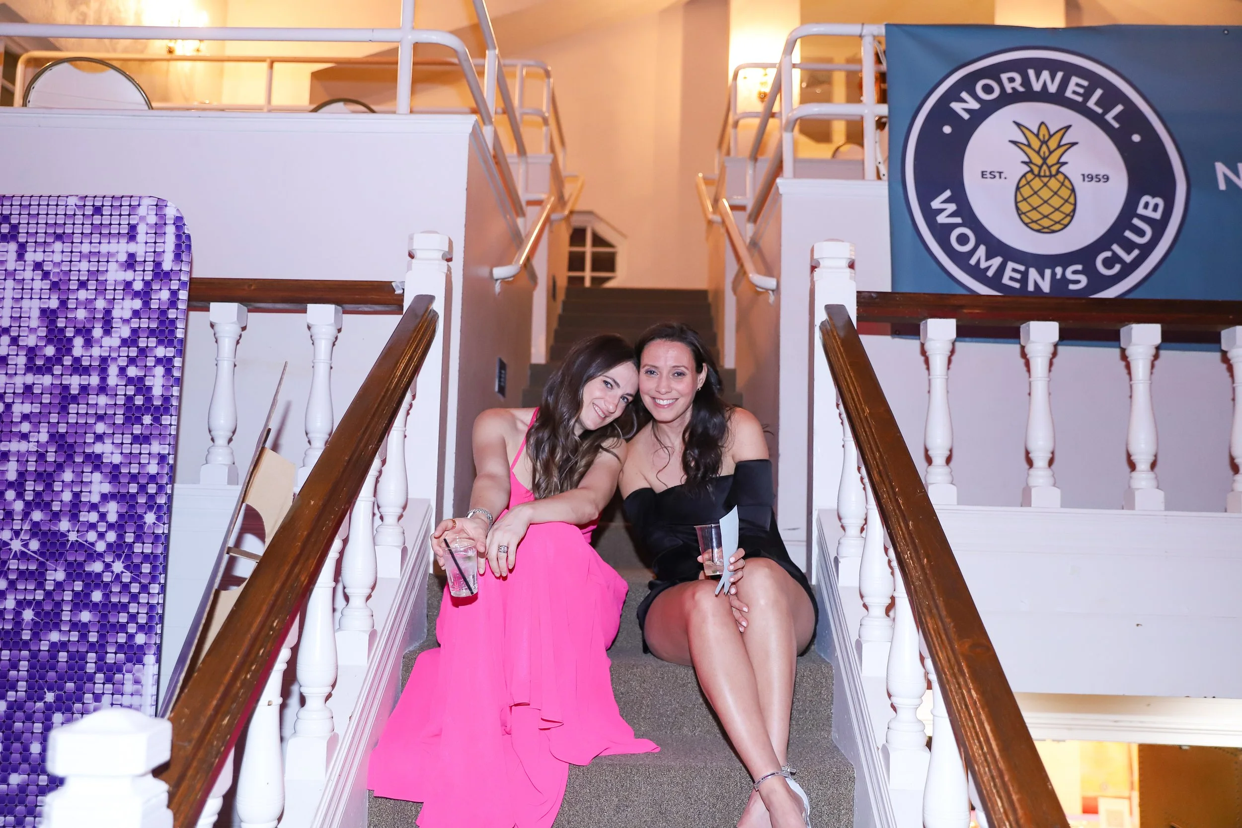 Two women sitting on a staircase inside a building, smiling and holding drinks, with a Norwell Women's Club banner hanging on the wall behind them.