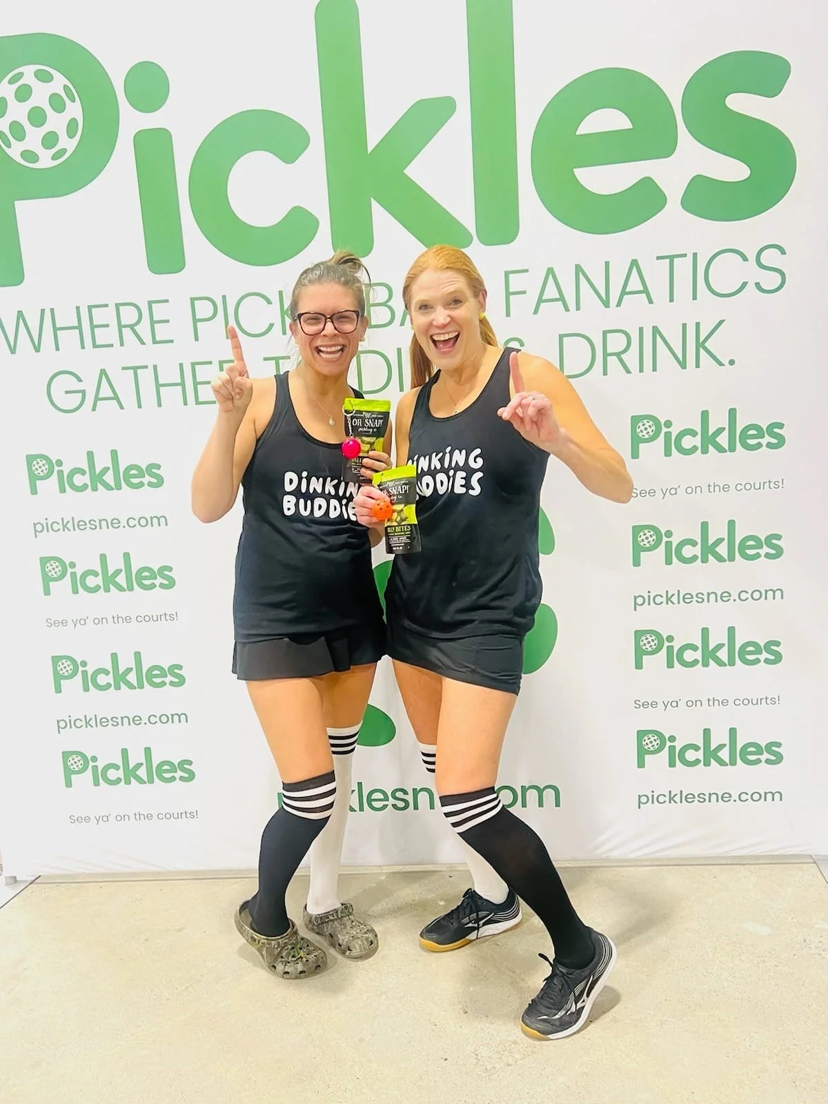 Two women in black athletic outfits and striped knee-high socks smiling and celebrating in front of a green and white Pickles backdrop at a pickleball event, holding snacks and wearing medals.
