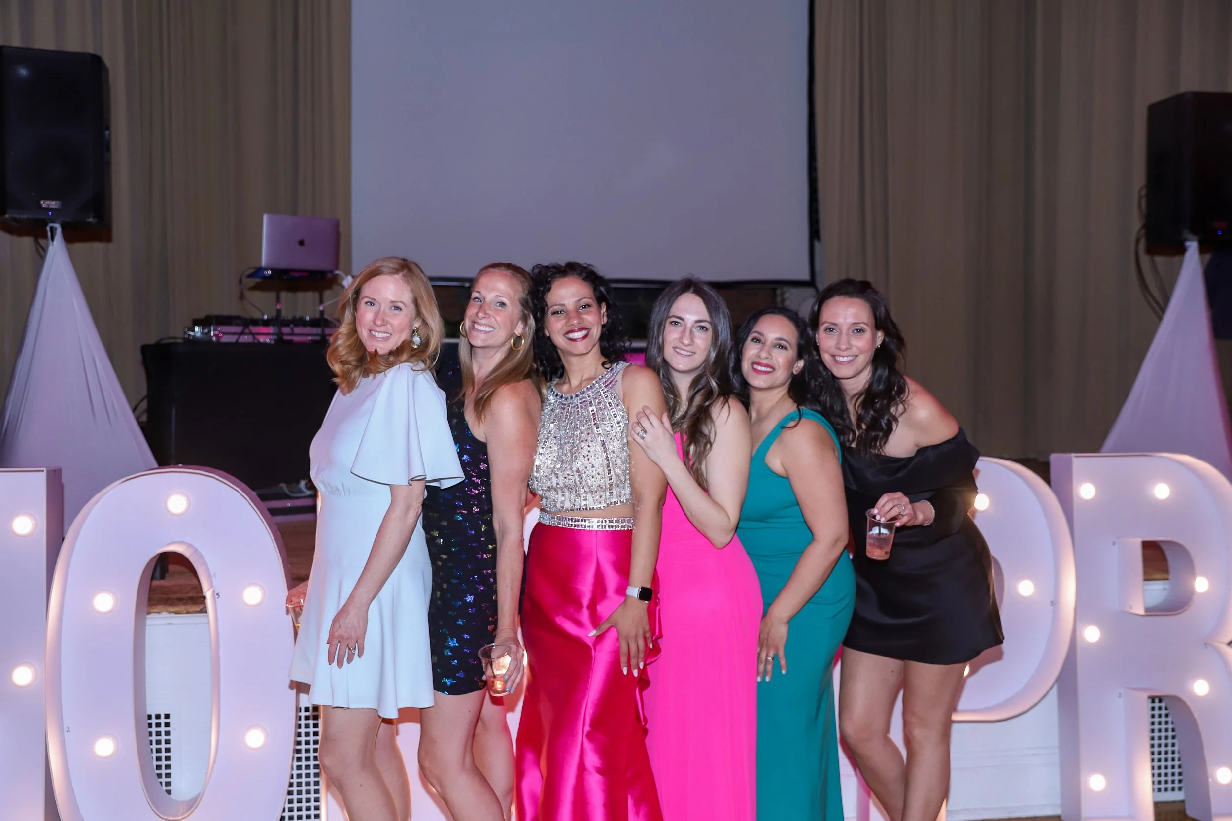 Six women in colorful dresses smiling at a party, standing in front of large illuminated letters spelling 'LOVE' at an indoor event with a stage and equipment in the background.