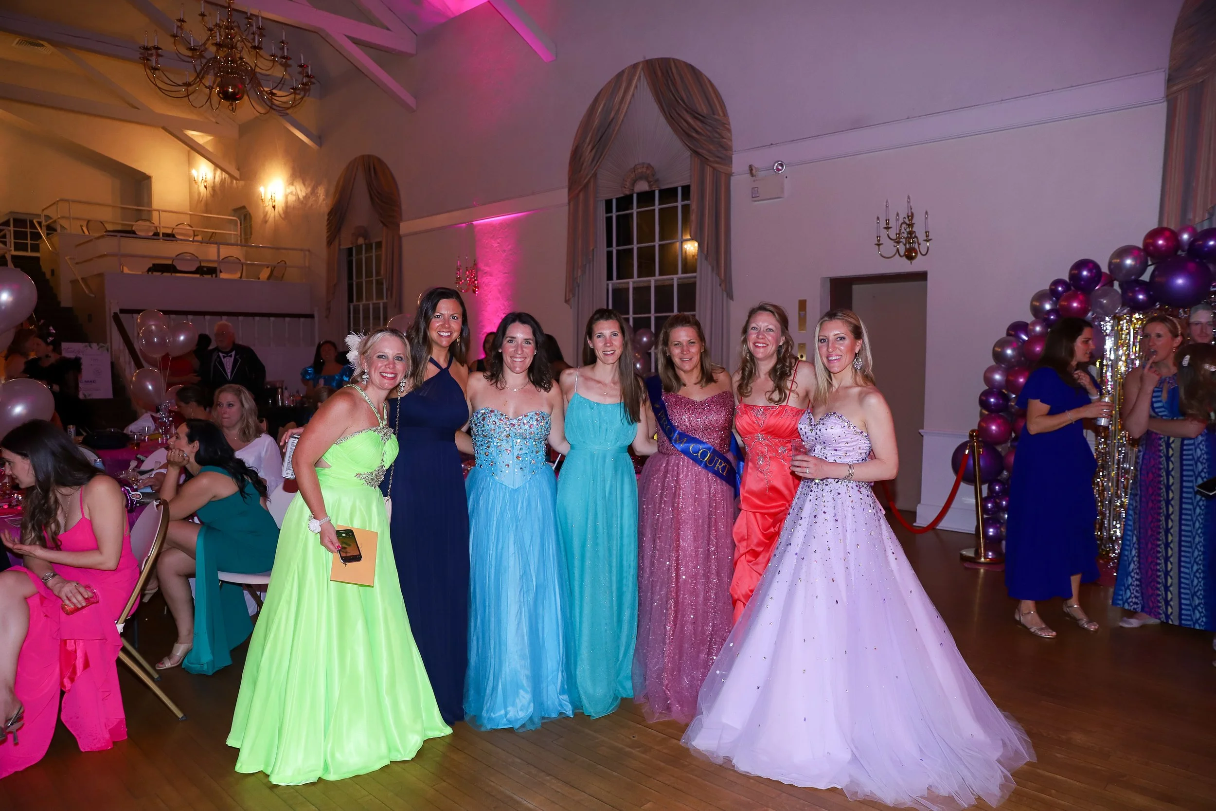 Women in colorful dresses posing at a formal event with balloons and decorations.