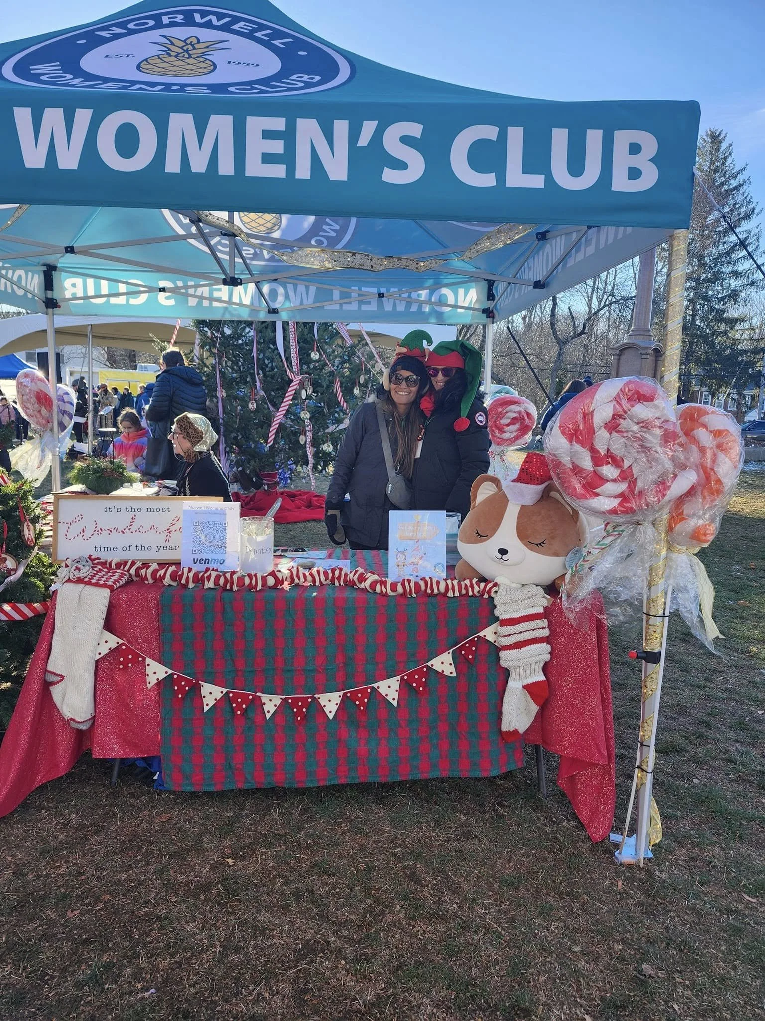 A booth under a blue tent labeled 'Women's Club' at a festive outdoor event. The booth is decorated with red and green plaid and Christmas-themed ornaments, featuring a stuffed dog wearing a Santa hat and a scarf, and large candy-themed decorations.