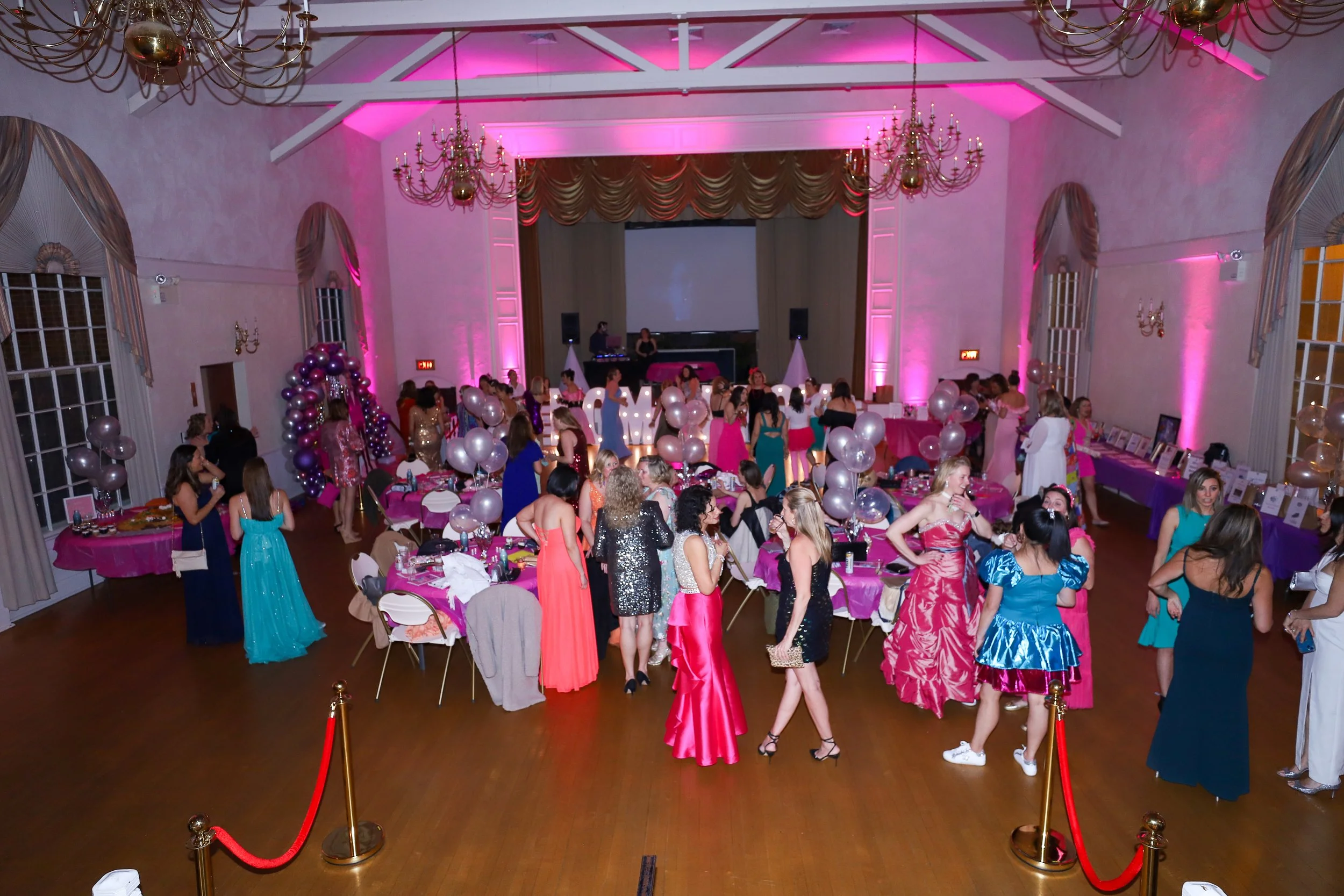 A large indoor party hall filled with people dressed in formal and semi-formal attire, with pink and purple lighting, balloon decorations, round tables covered with pink tablecloths, a stage at the front with a large projector screen, and a DJ setup.