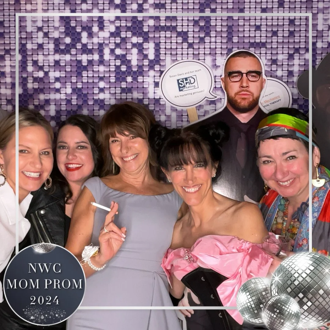 A group of six women and one man posing together at a prom event with a purple mosaic backdrop. Some women are smiling and holding drinks, one woman is holding a cigarette. The group is celebrating NWC Mom Prom 2024, with festive decorations like dis