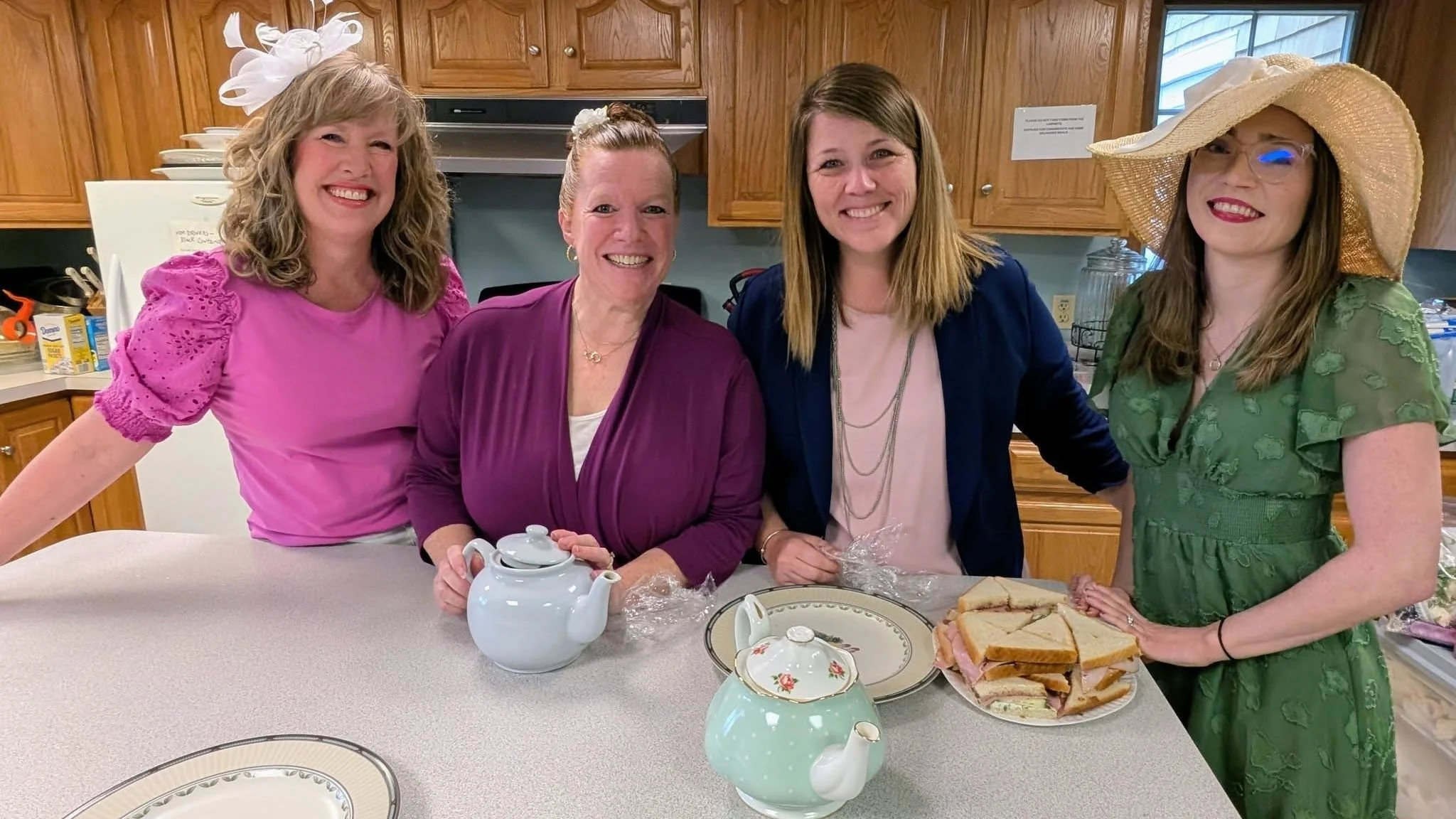 Four women standing together in a kitchen, smiling at the camera. The woman on the right is holding a plate of sandwiches, and there are teapots on the counter in front of them.