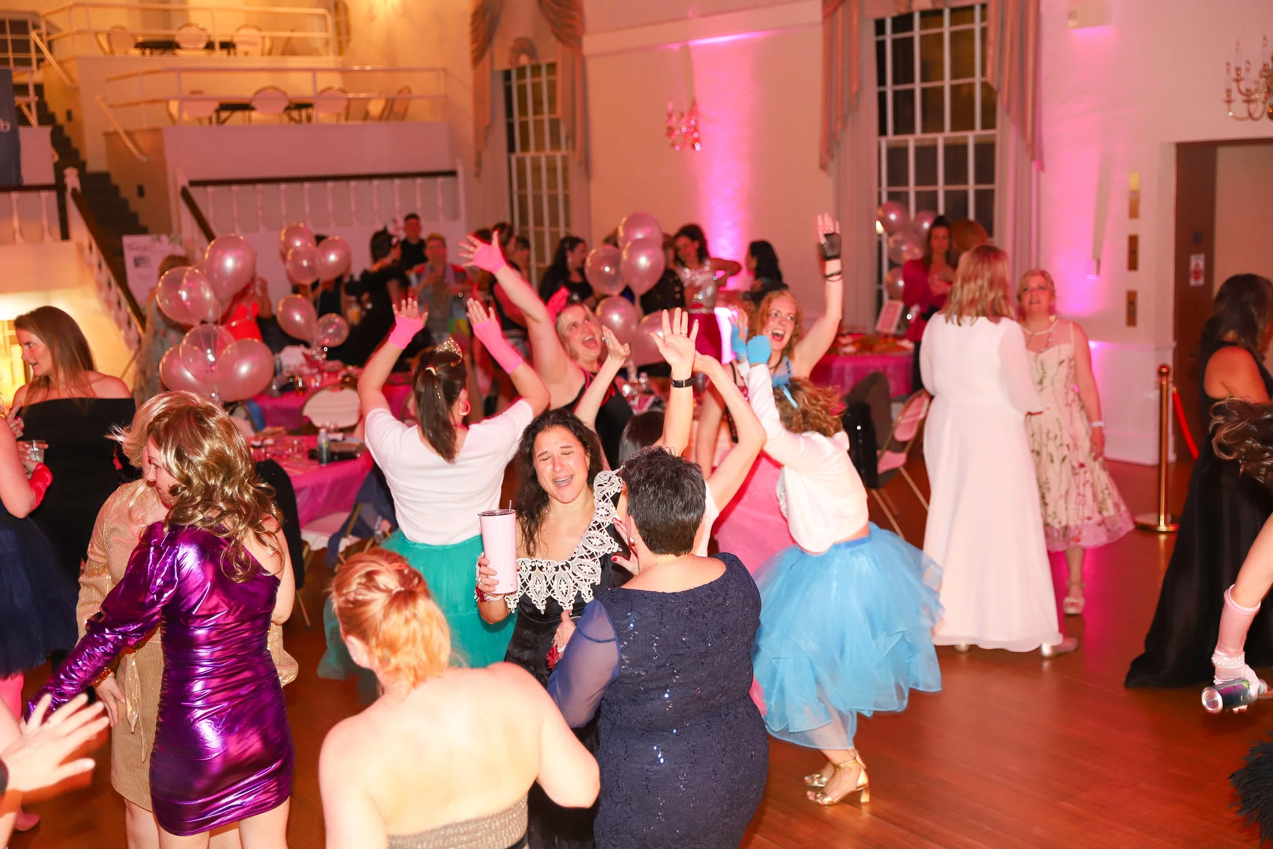 People dancing and celebrating at a party with pink balloons and pink lighting.
