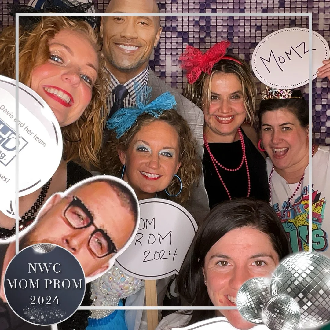 A group of women and a man at a mom prom event, holding signs with messages like "Momz" and "Mom from 2024". They are dressed festively with colorful accessories and smiling for a photo.