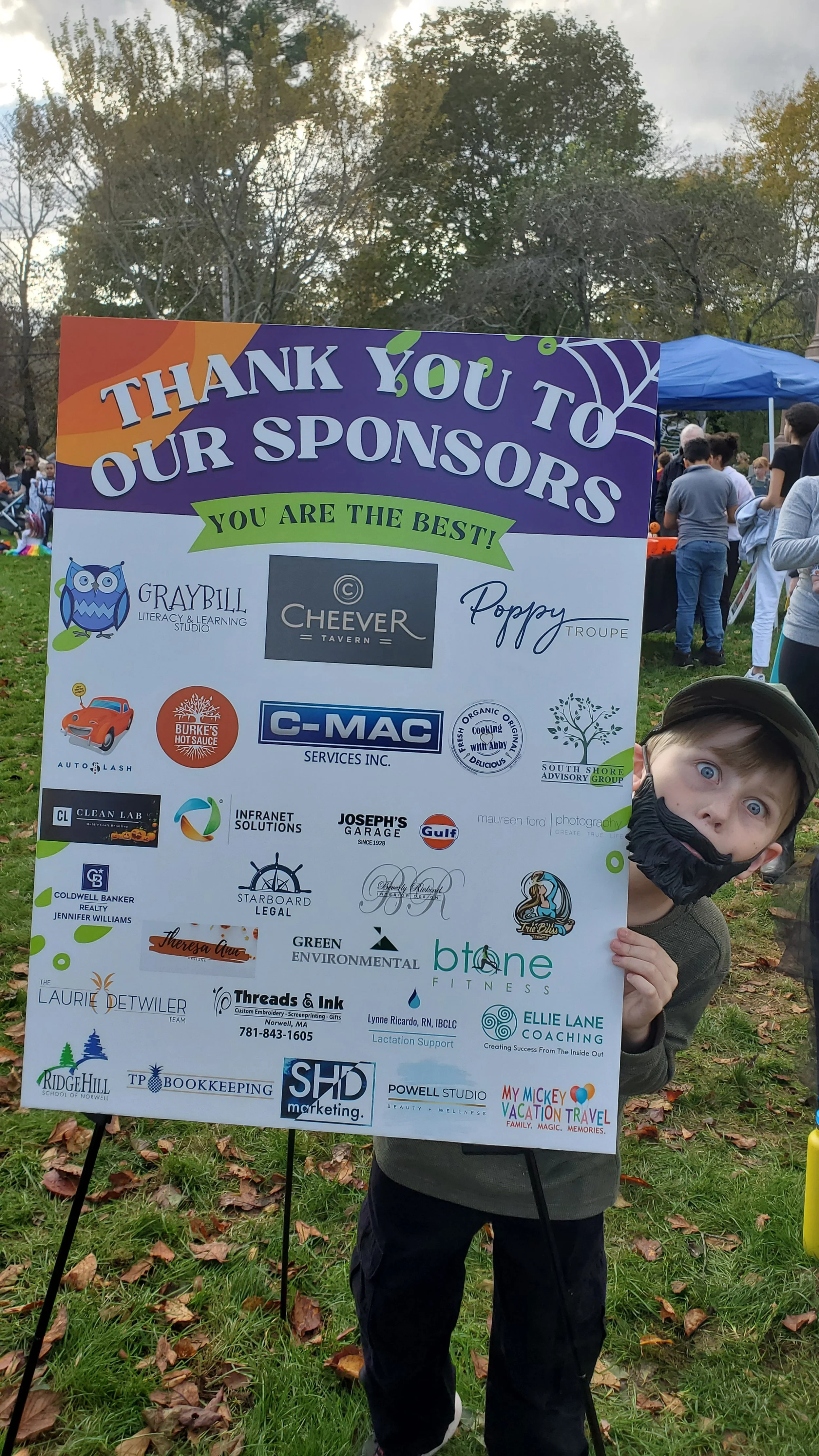 A boy with wide eyes and a black face mask holding a sponsorship board at an outdoor event with trees and tents in the background.